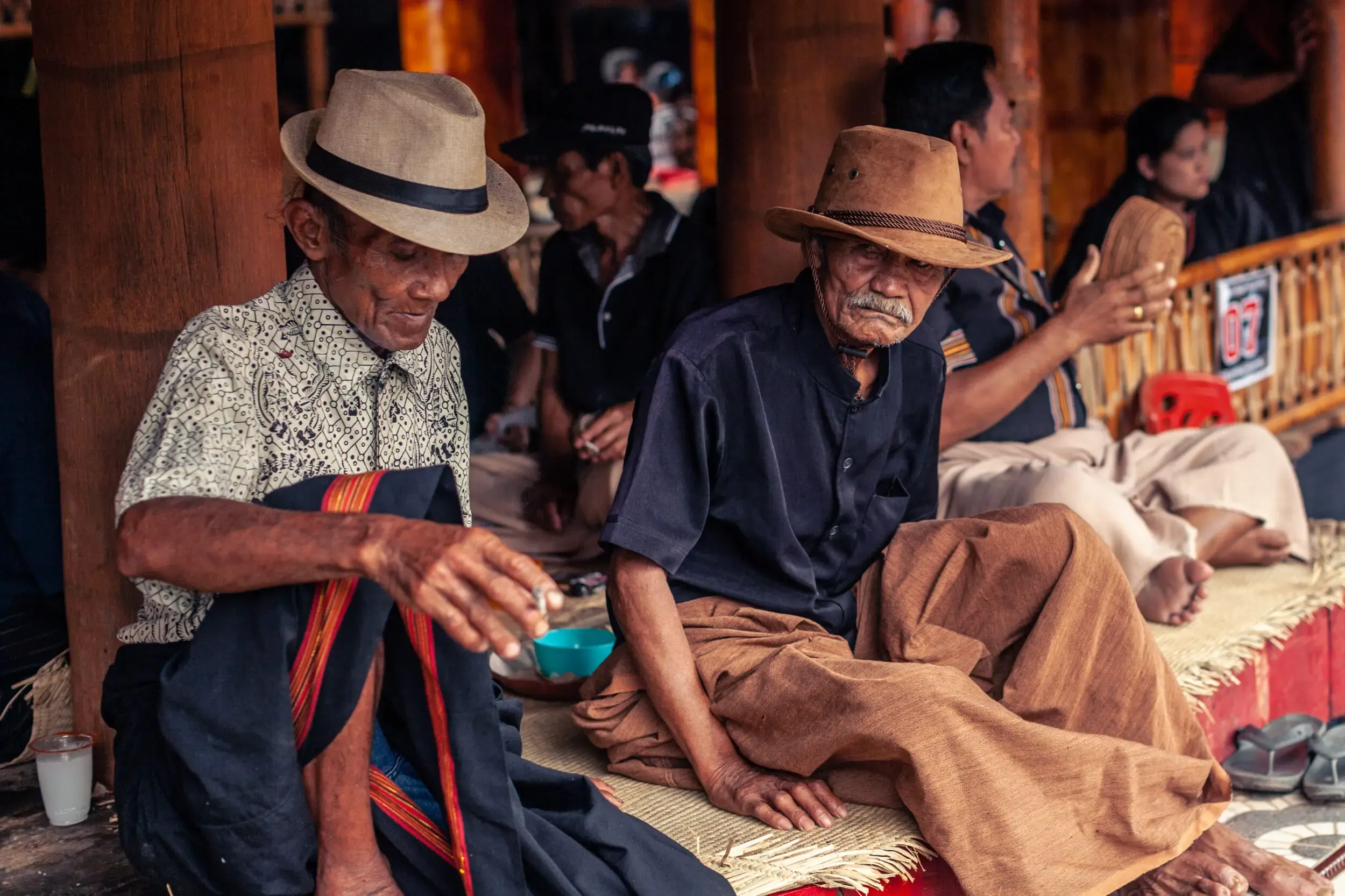 Two elderly men wearing traditional hats and clothing, seated on a mat in a cultural setting.