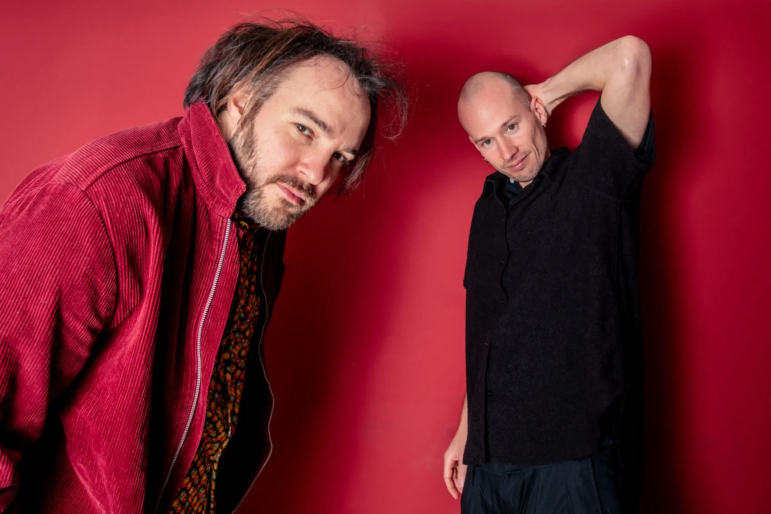 Two men posing against a red background, one in a red corduroy jacket and the other in a black shirt.