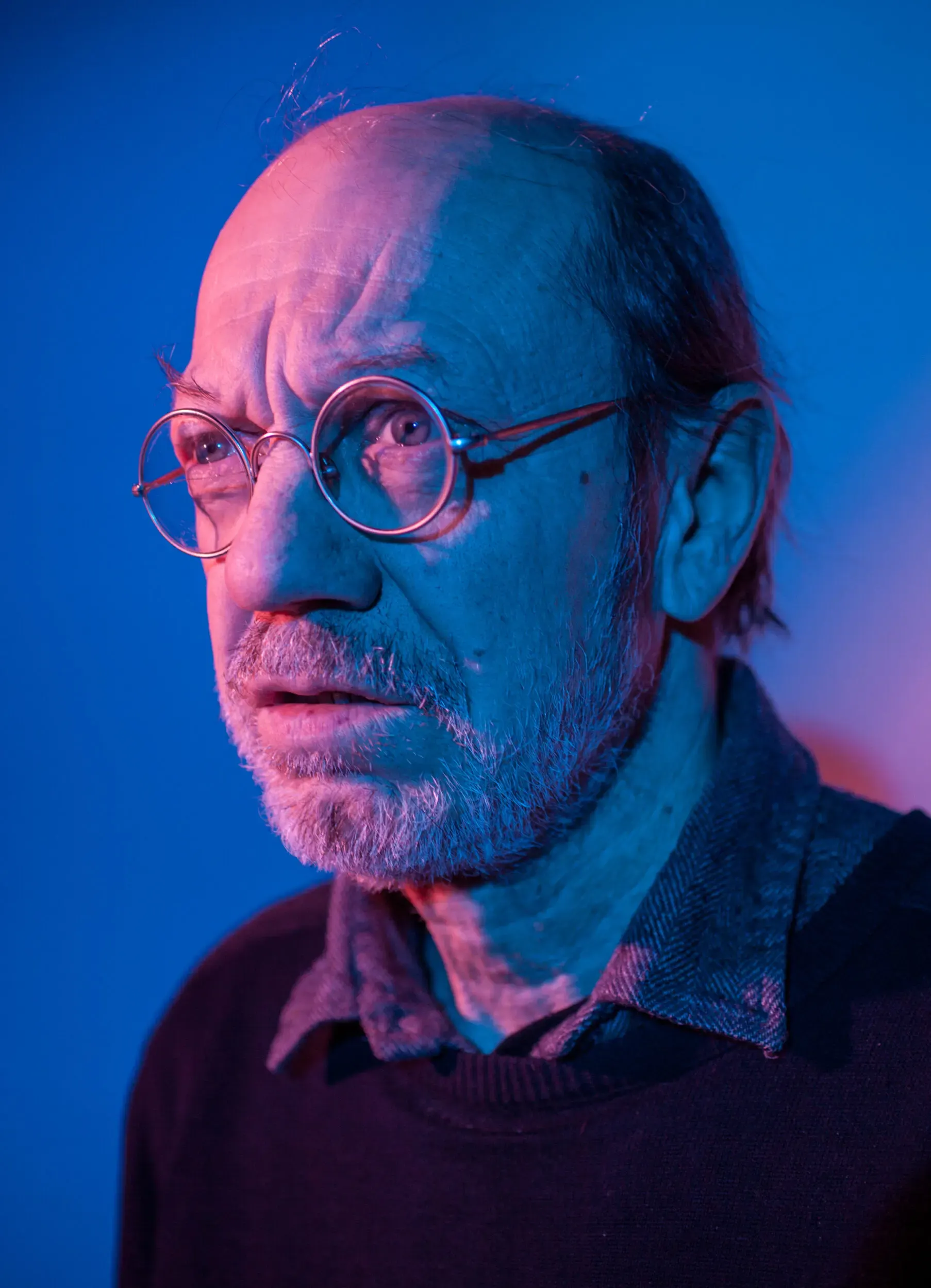 A man with glasses and a beard, wearing a dark sweater, illuminated by blue and purple lighting.