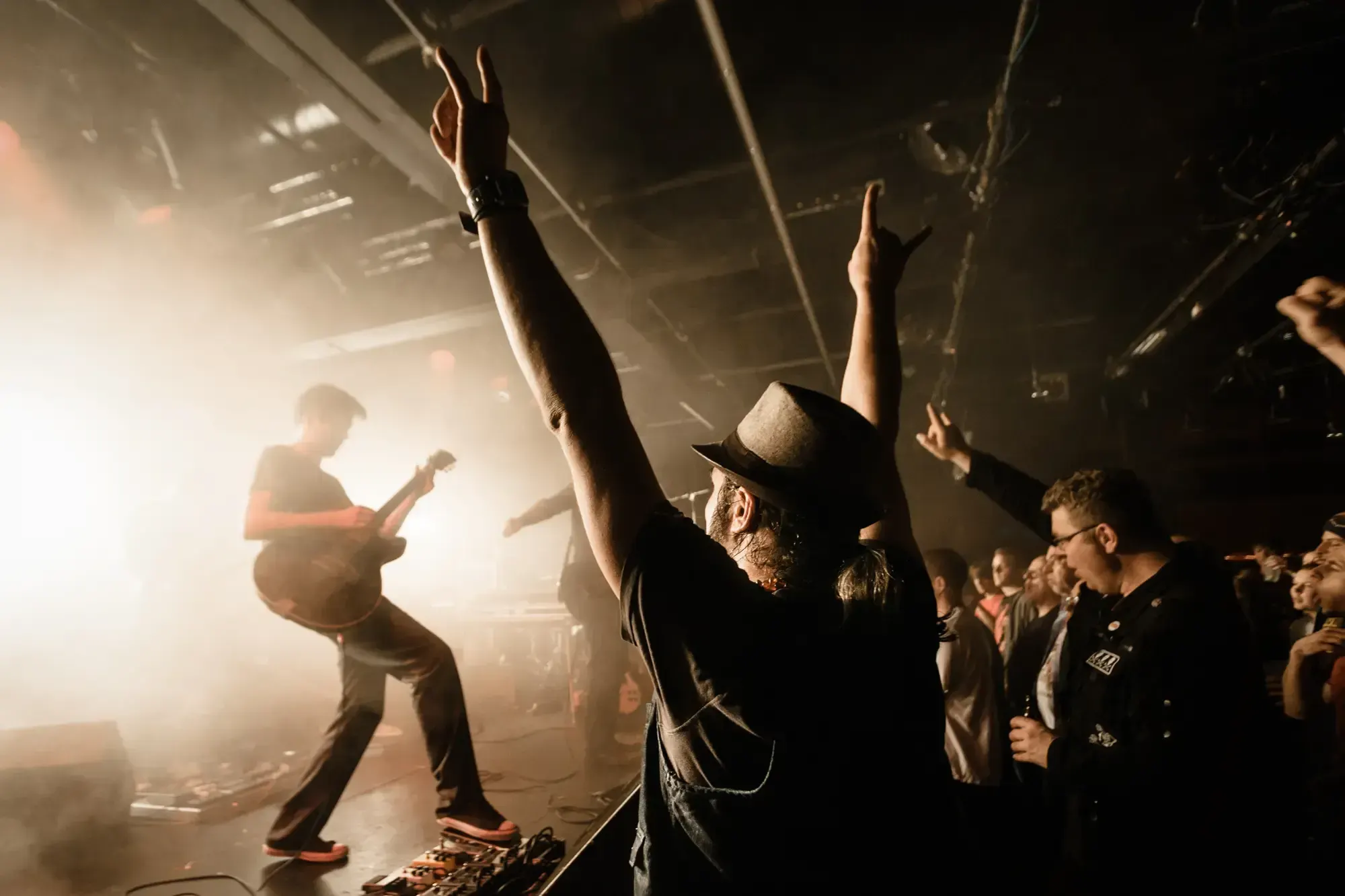 Fans raising their arms and cheering in front of the stage at a concert, a guitarist performing in the background.