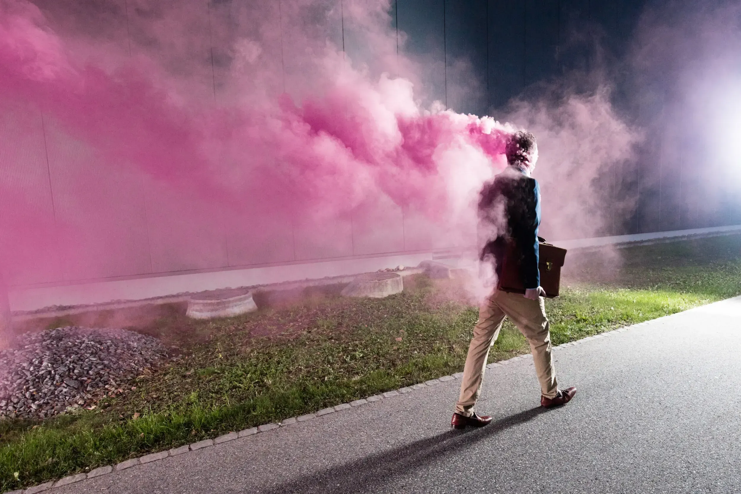 Man in a suit walking down a path at night trailing pink smoke from a smoke bomb, briefcase in hand.