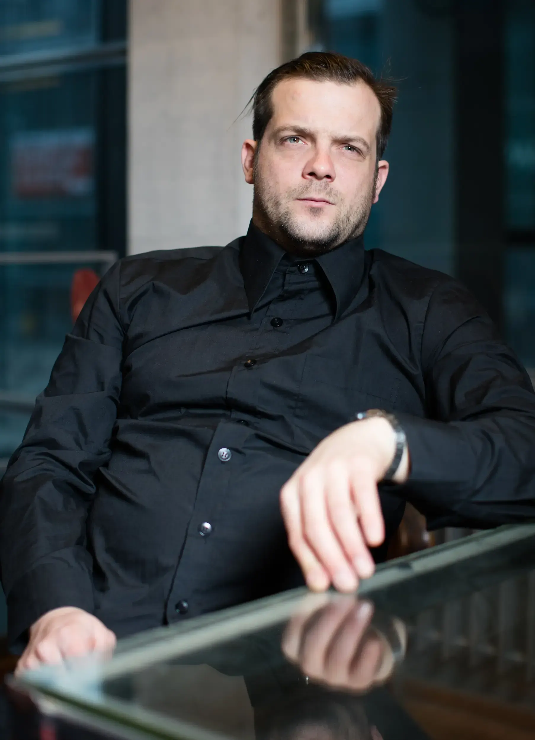André Willmund in a black shirt resting his arm on a reflective glass table by a window.