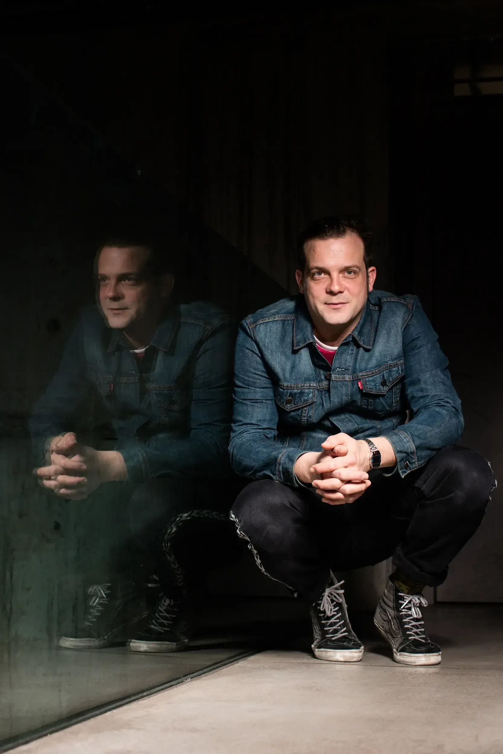 André Willmund crouching beside a glass wall that reflects his image, hands clasped.