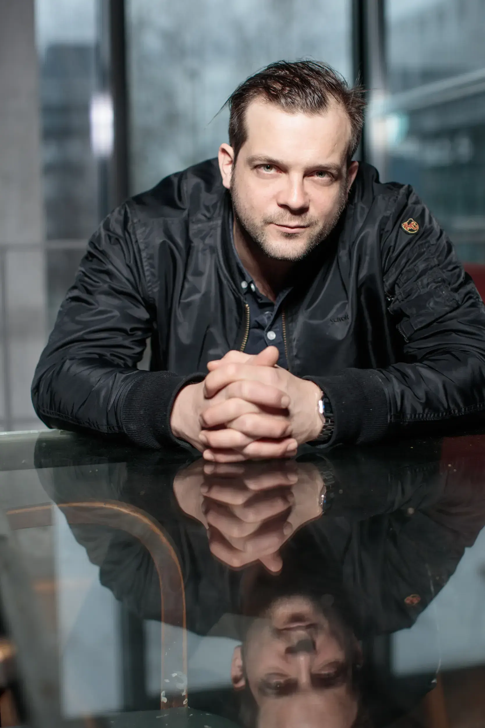 André Willmund leaning on a reflective glass table with hands folded, window backdrop behind him.
