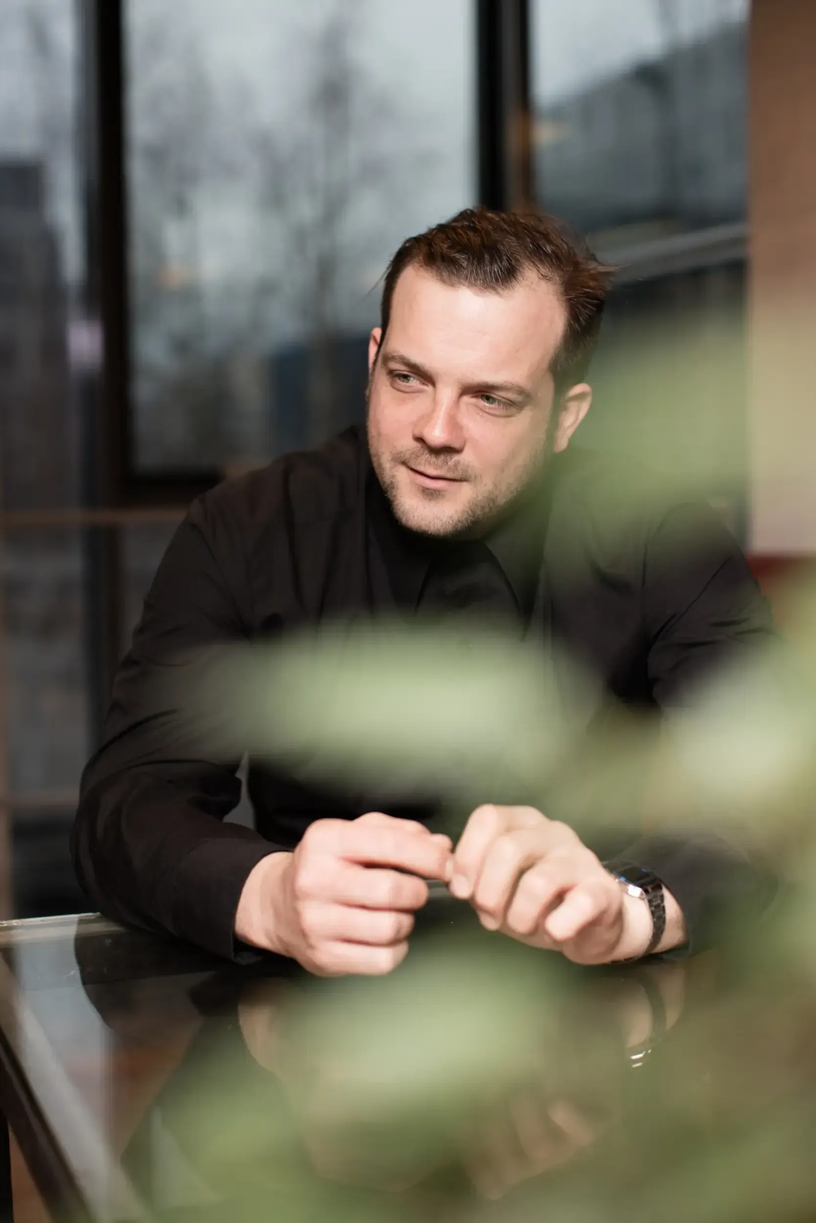 Smiling portrait of André Willmund seated at a glass table with green plants blurred in the foreground.