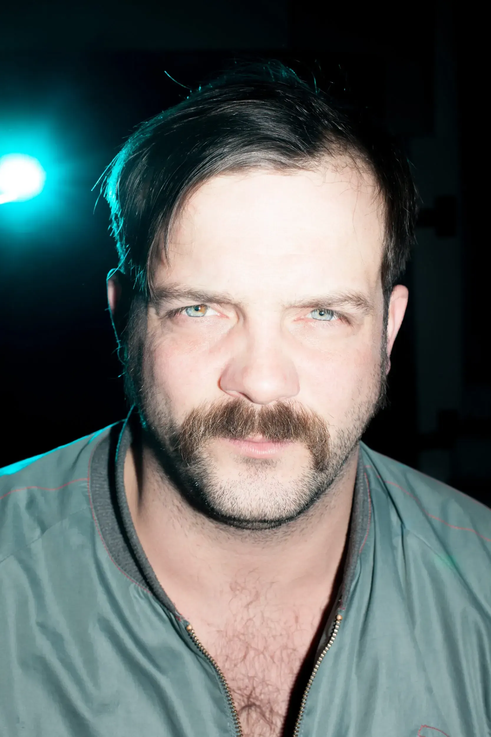 Portrait of André Willmund with a moustache, lit by a single teal light against a dark background.