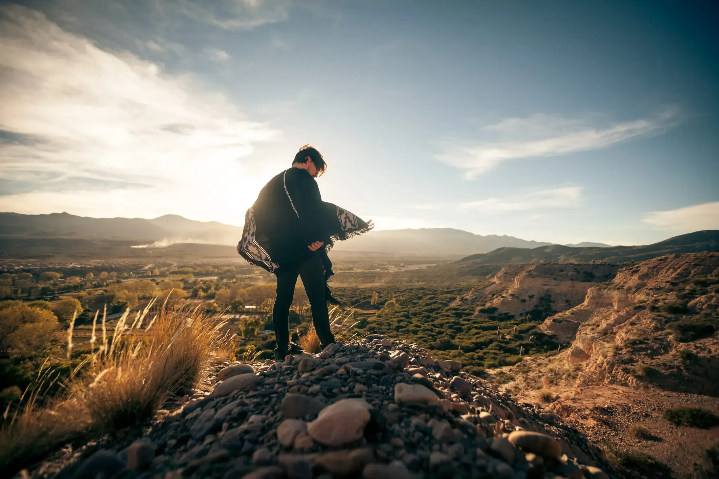 Man wrapped in a poncho standing on a rocky ridge overlooking a valley in Argentina at sunset.