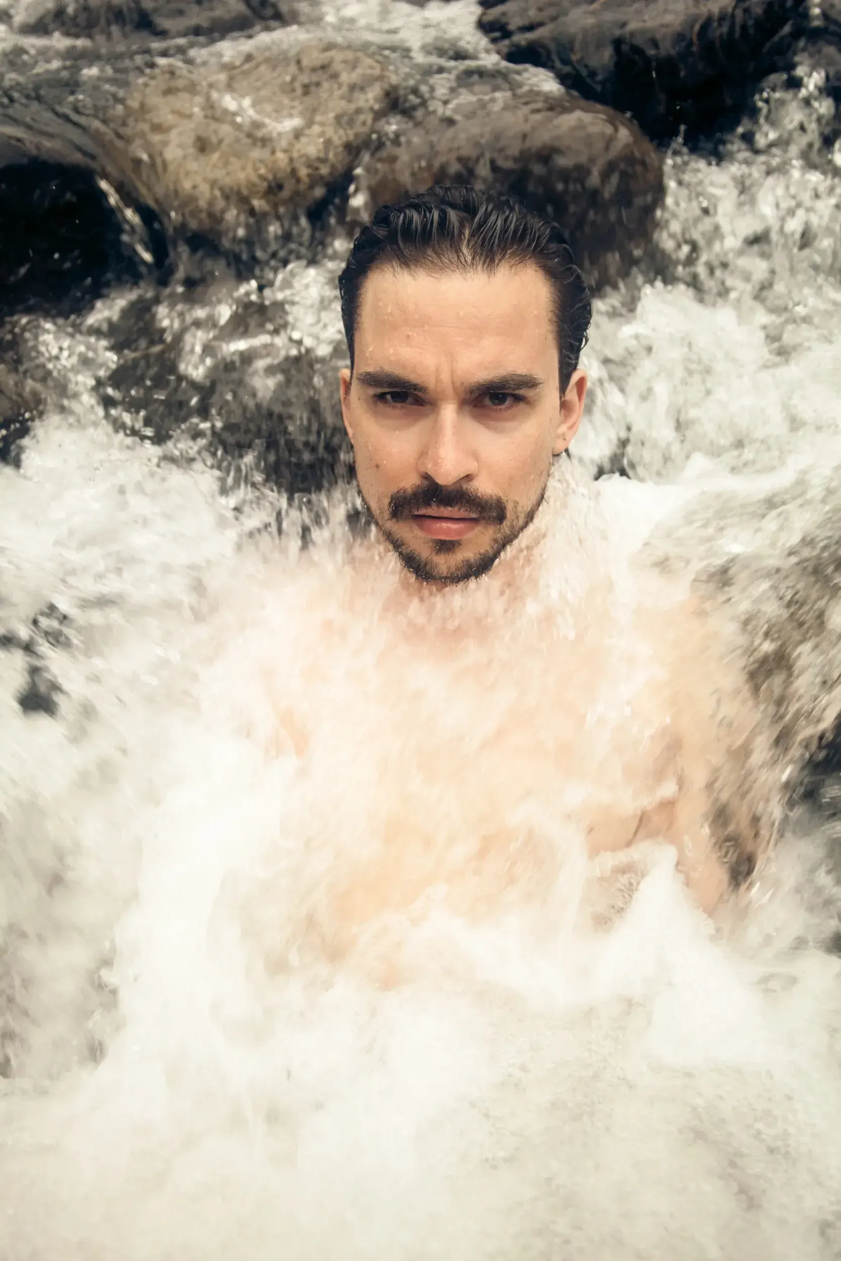 Man's face emerging from a rushing waterfall, water crashing over his shoulders.