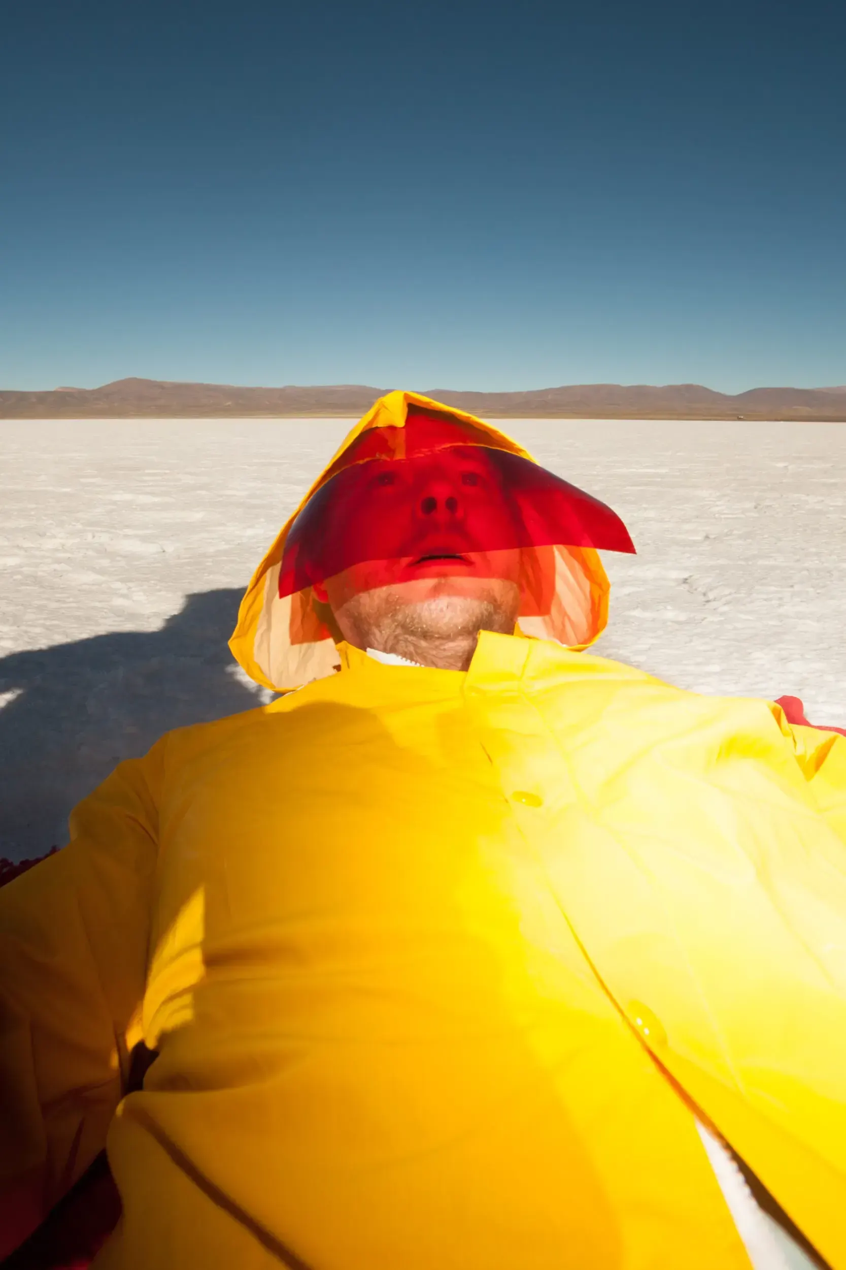 Close-up of a man in a yellow raincoat with a red-lit face, lying on the white salt flats of Argentina.