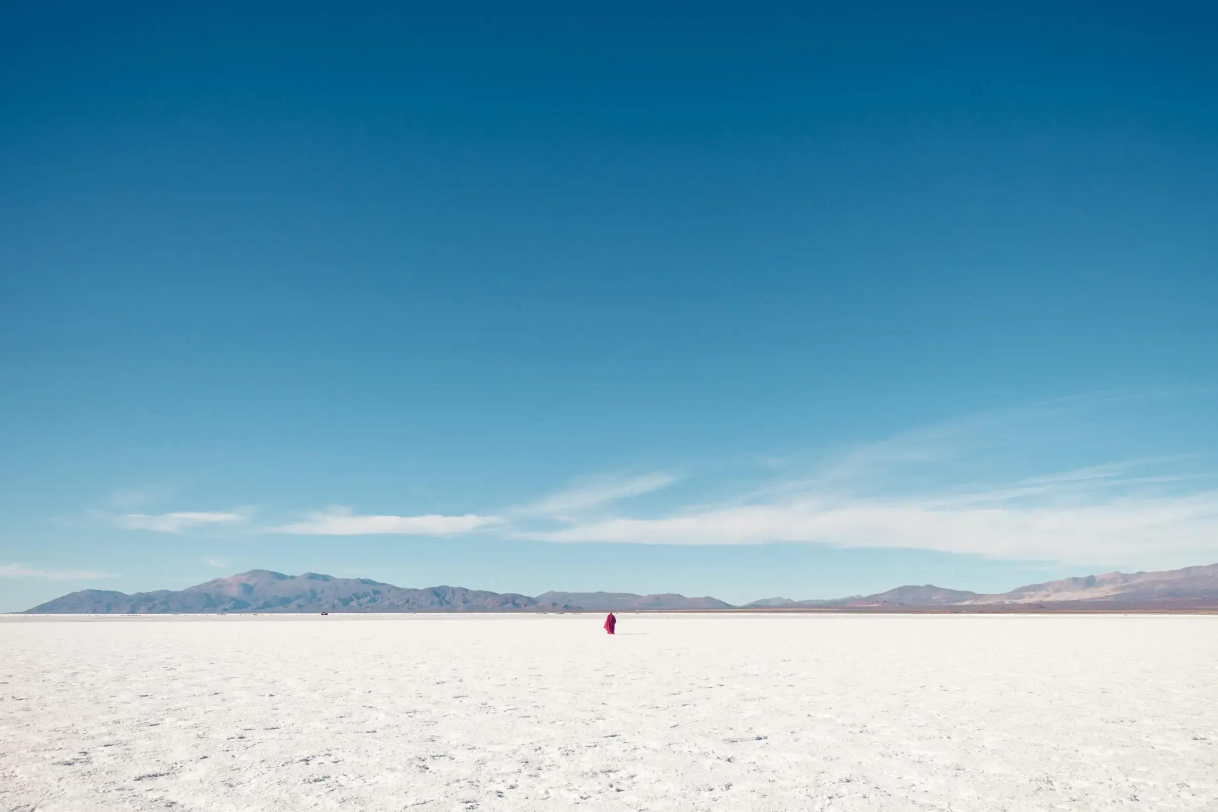 Small red-clad figure standing alone on the vast white salt flats of Argentina beneath distant mountains.
