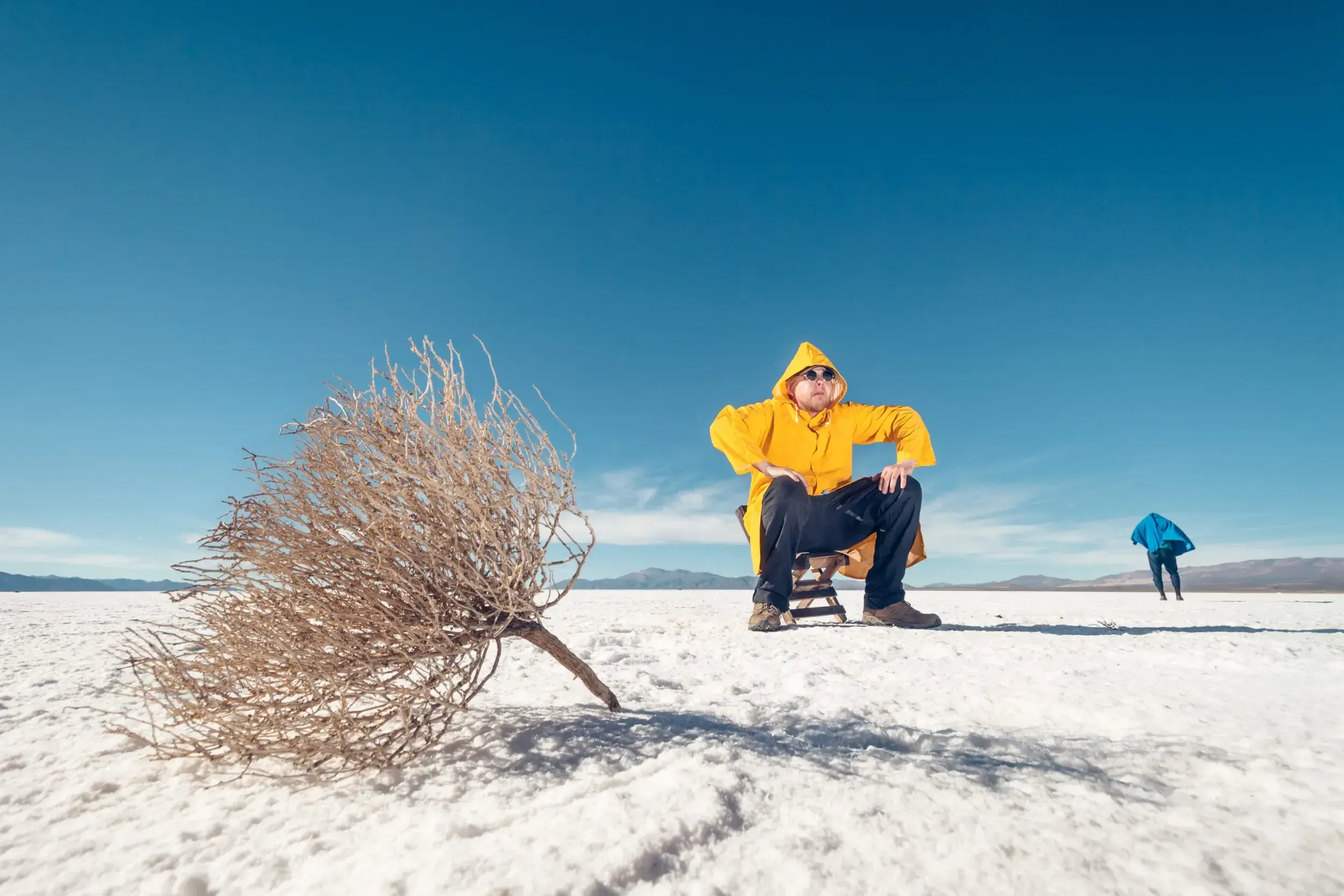 Man in a yellow raincoat sitting on a stool in the white salt flats of Argentina, a tumbleweed nearby.