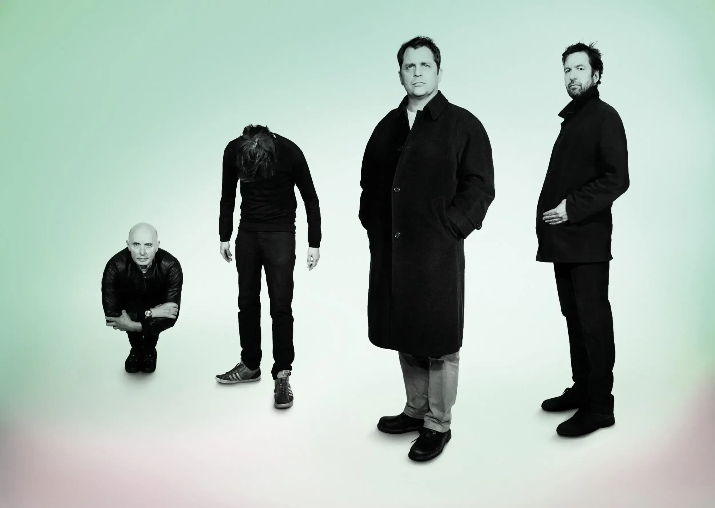 Theatre ensemble recreating a Stranglers band photo, four men posing at different heights in black coats.