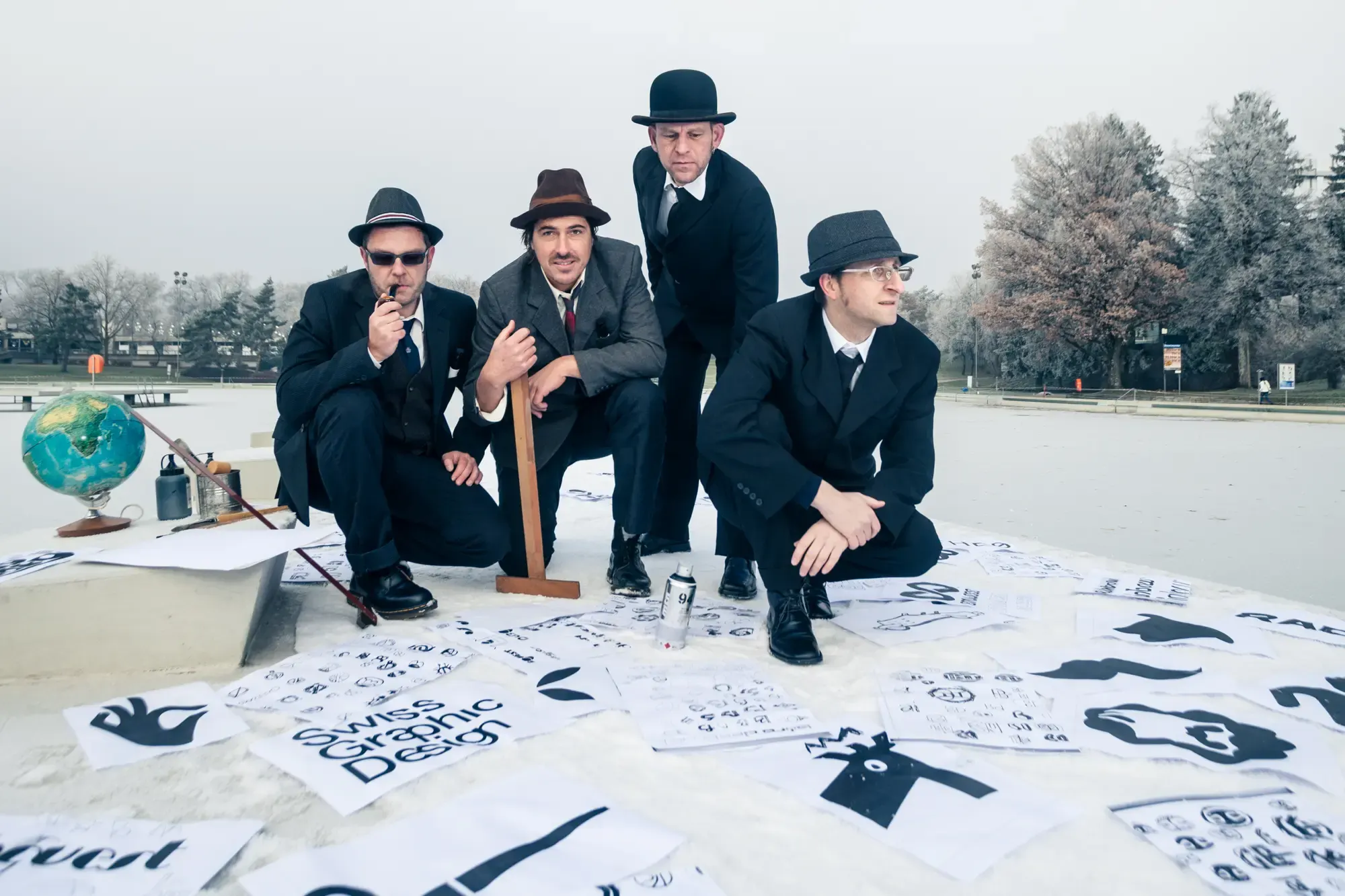 Four men in suits and hats, surrounded by design papers, a globe, and a ruler on a snowy surface.