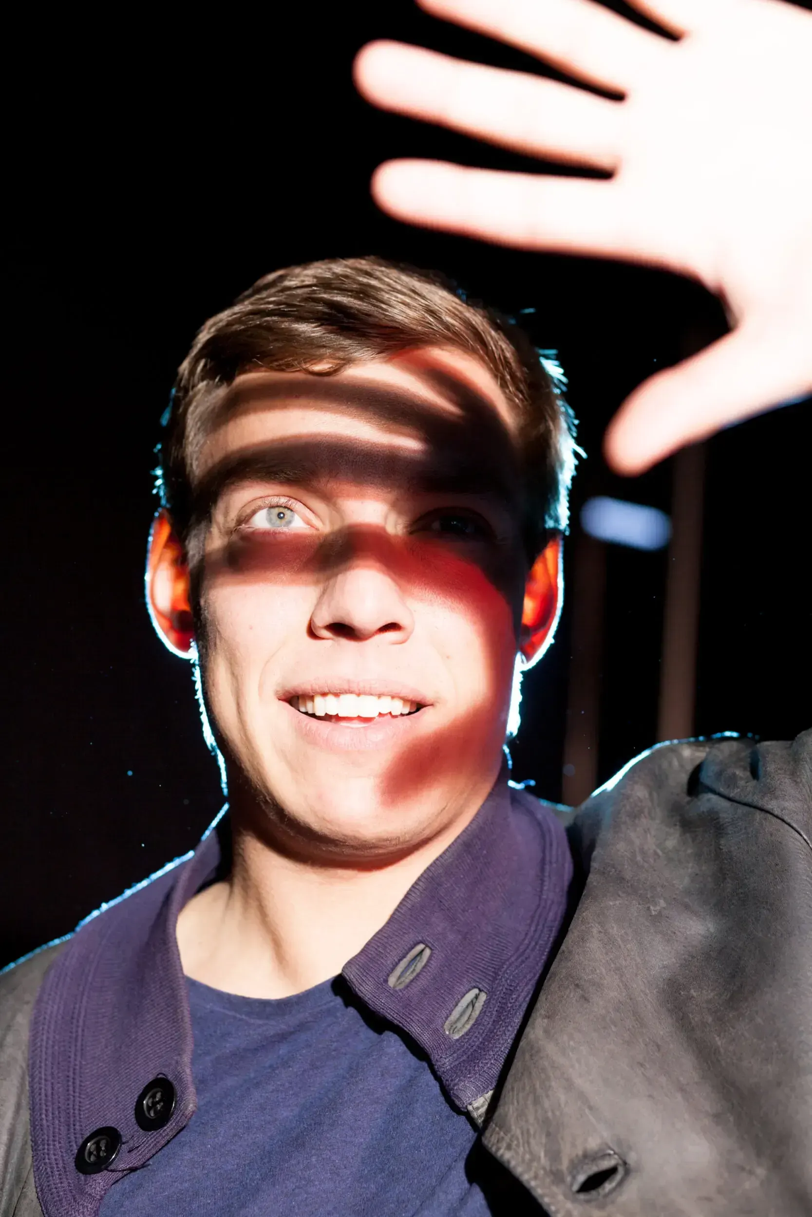 Close-up portrait of actor Benedict Fellmer smiling, with a hand casting a striped shadow across his face.