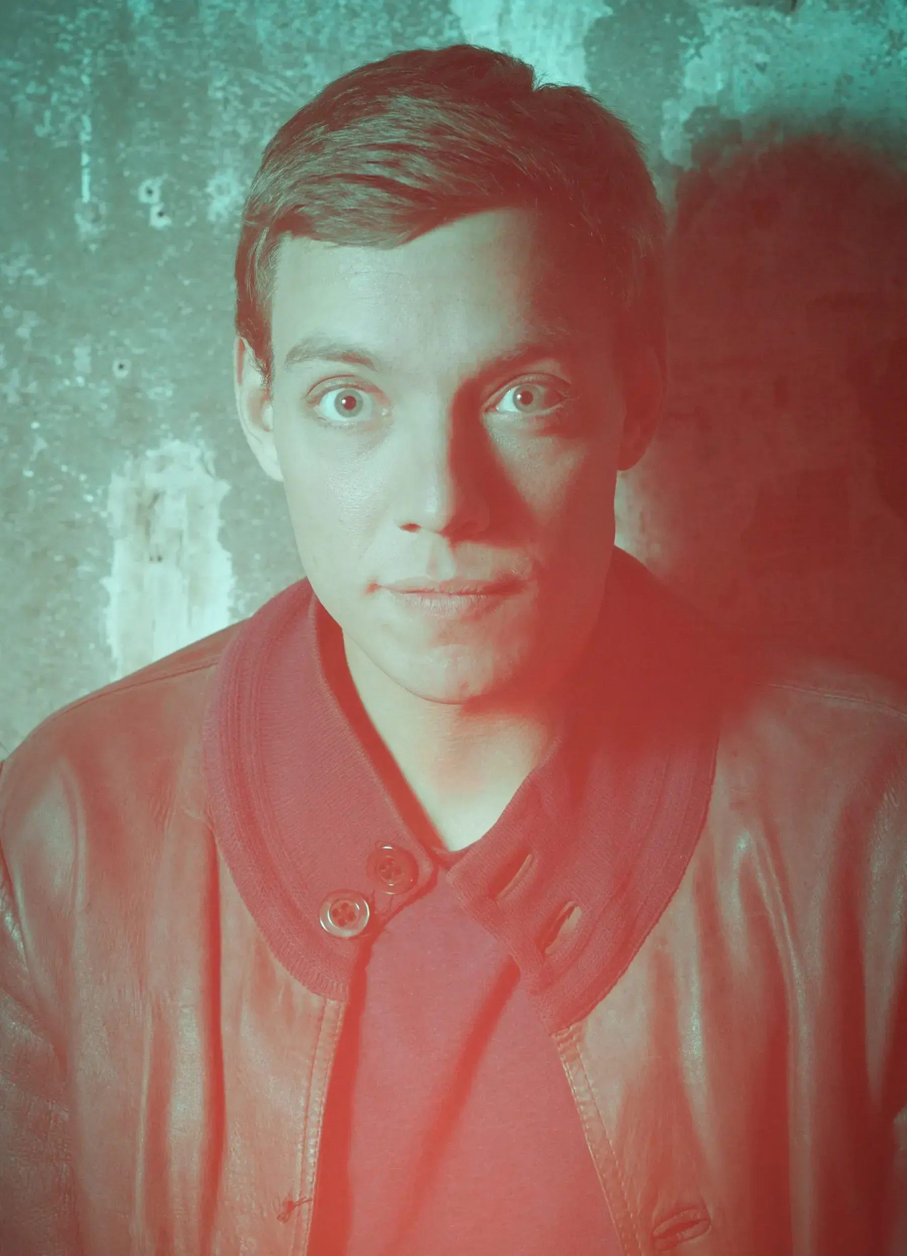 Portrait of actor Benedict Fellmer in a red leather jacket, lit in red and teal against a weathered wall.