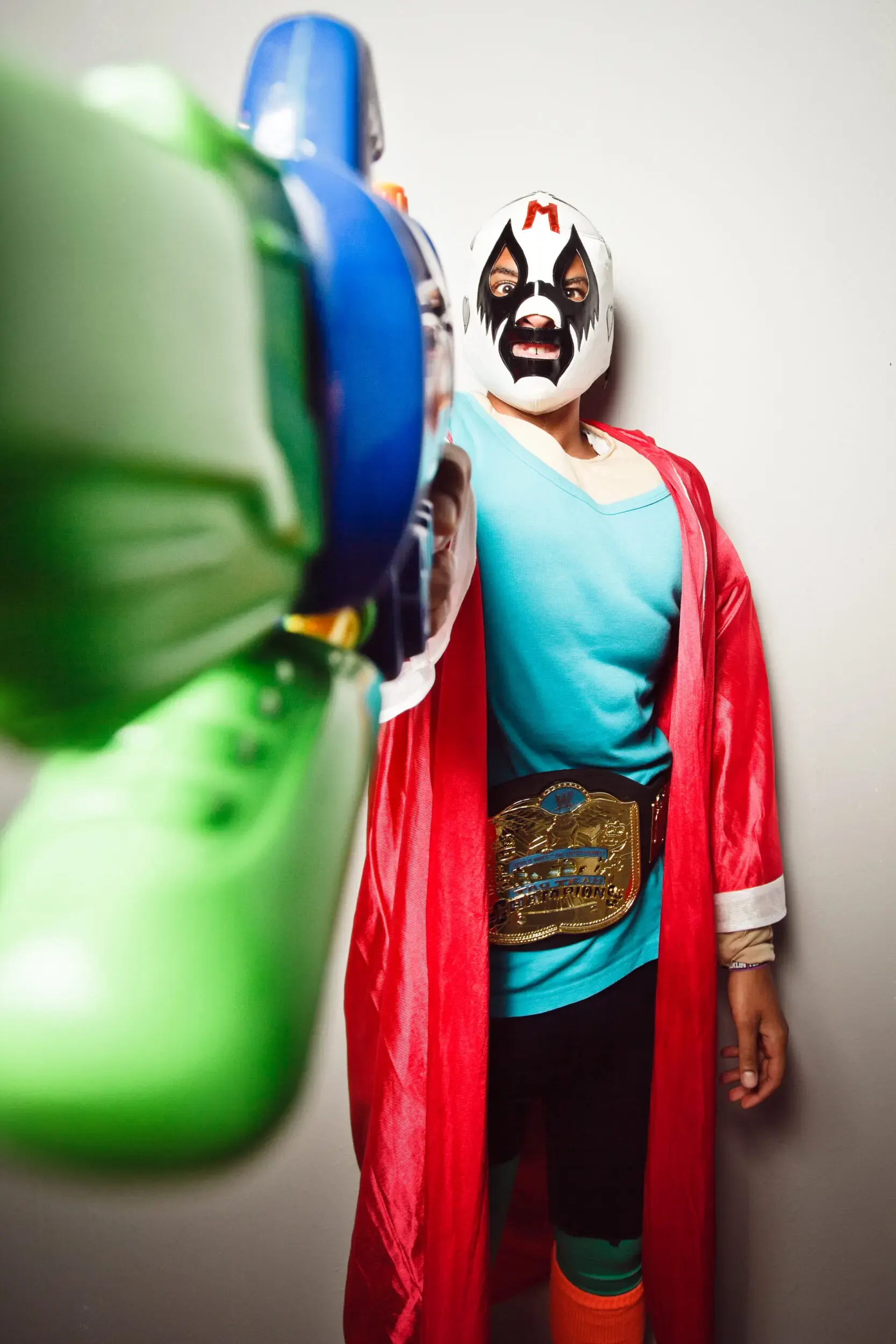 A wrestler in a red cape holds a green toy gun, wearing a white mask and a championship belt, with a blue shirt.