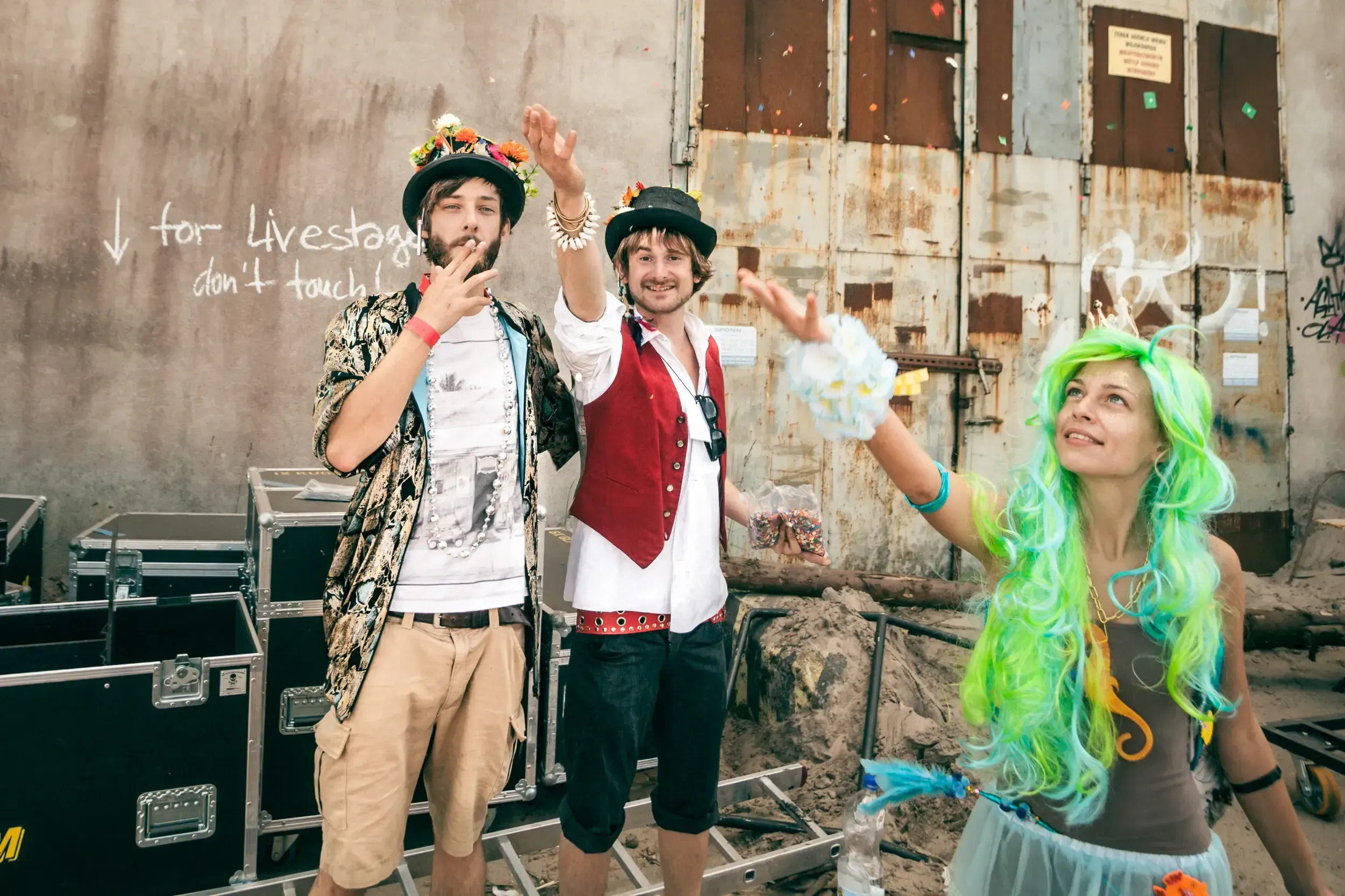 Three festival crew members backstage throwing confetti in front of a graffiti-covered wall reading 'for livestage don't touch'.