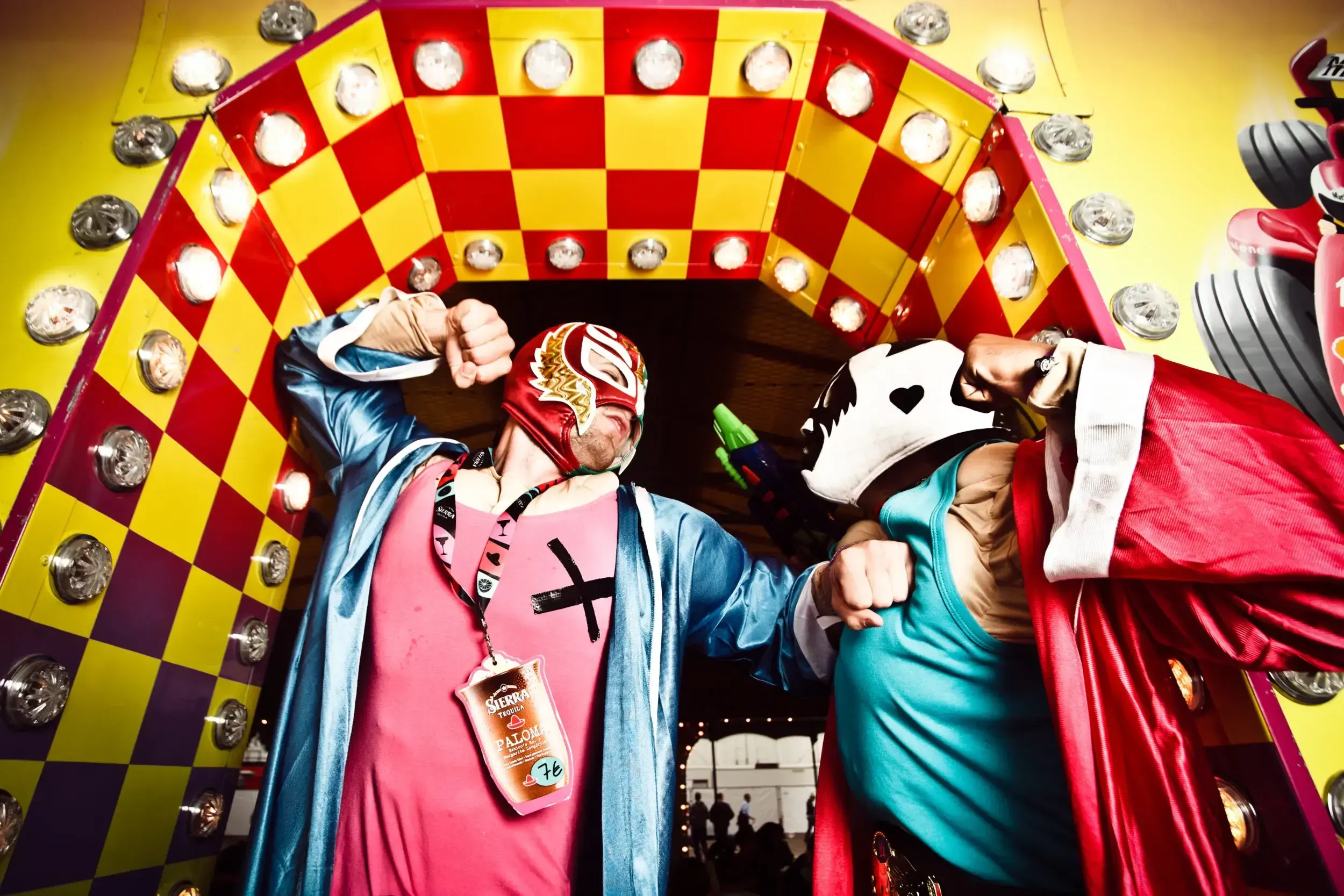 Two costumed festival-goers in wrestling masks and capes bump fists in front of a lit-up carnival ride.