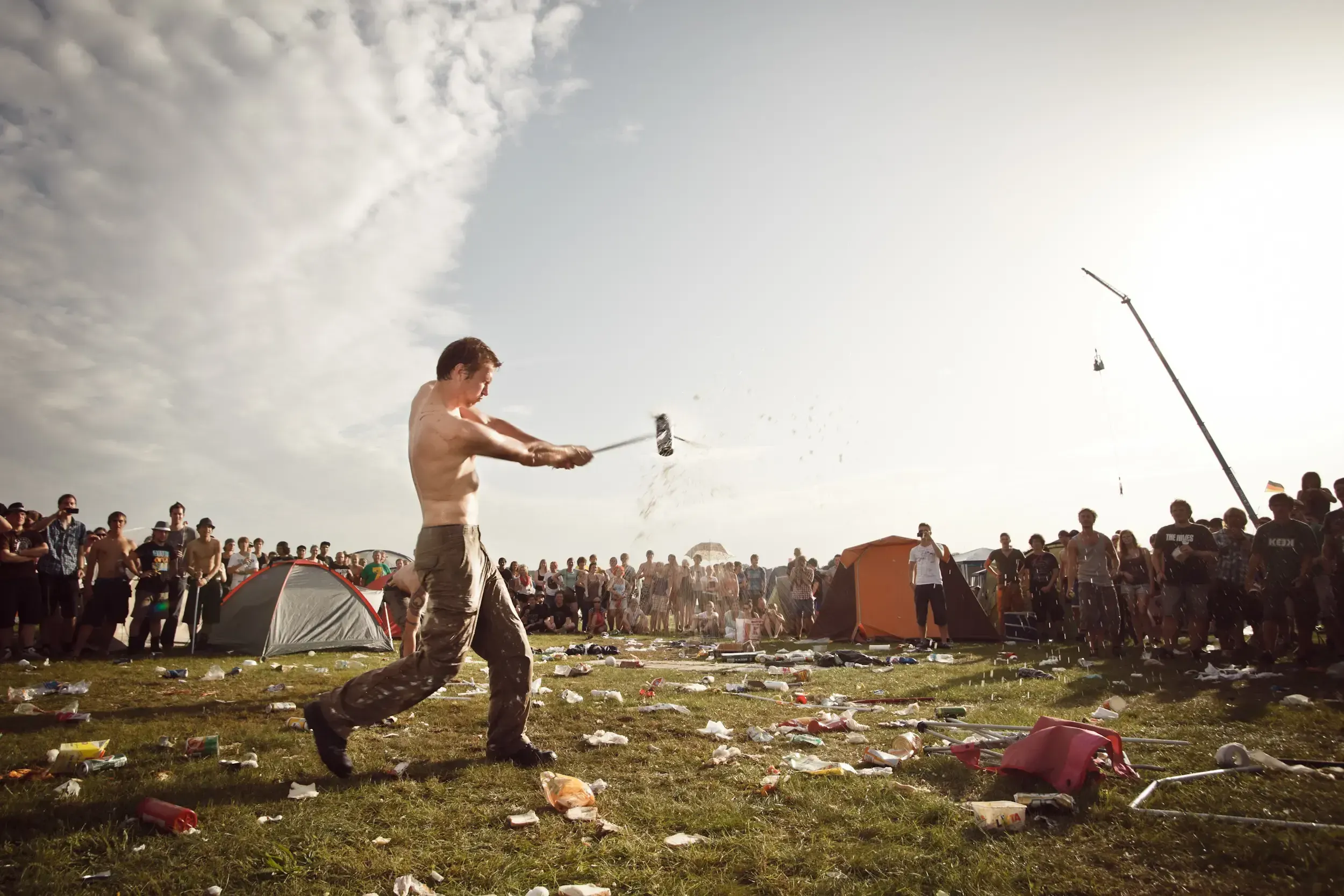 Shirtless man swinging a mallet at a high-striker strength game on a littered festival field.