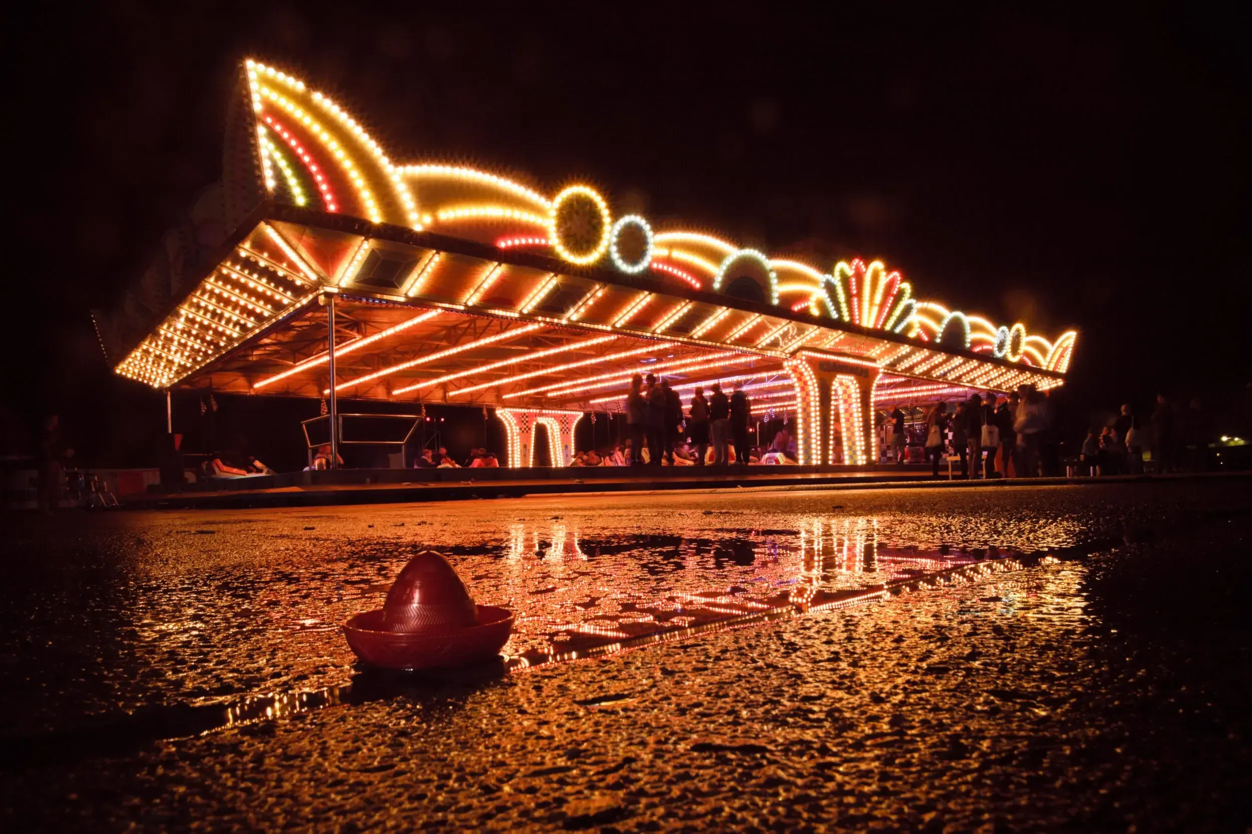 Illuminated fairground ride glowing at night, reflected in a puddle with a sombrero hat in the foreground.