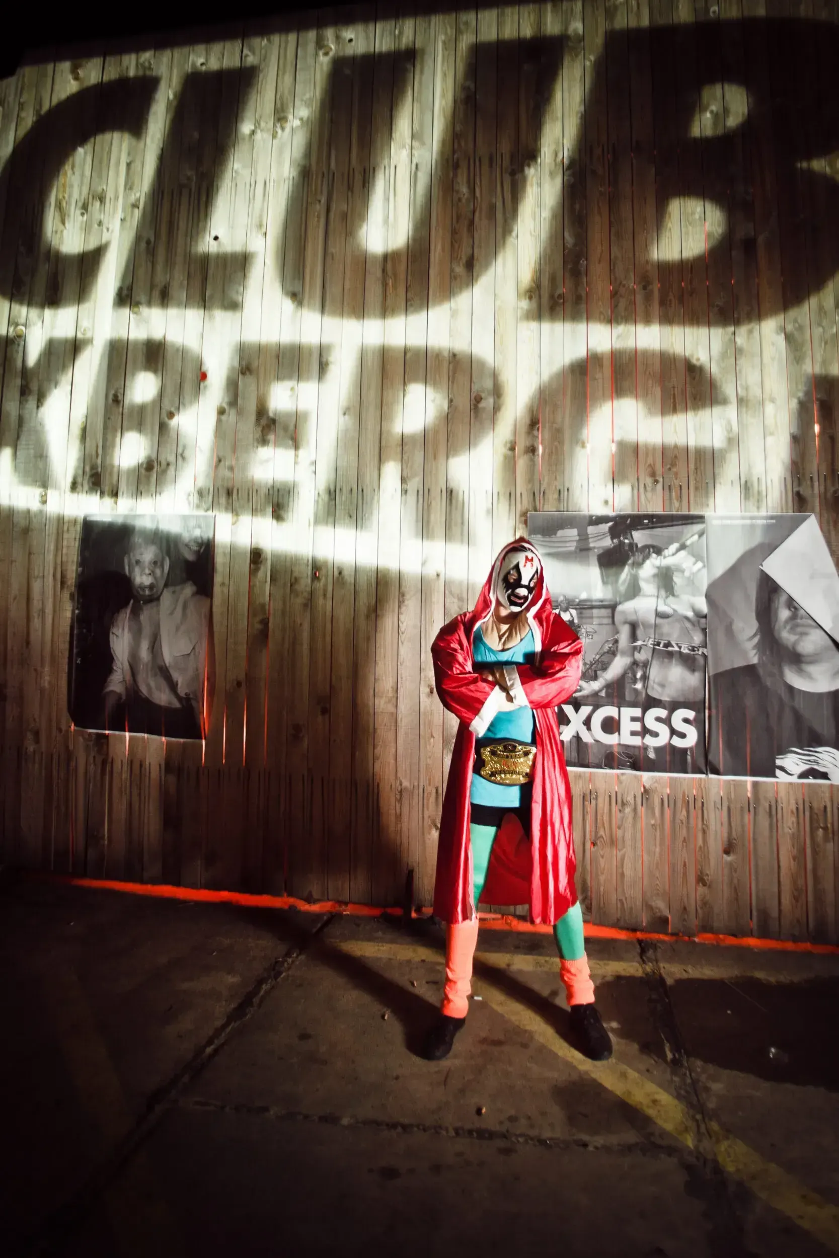 Person in a lucha libre mask and red robe posing in front of a wooden 'Club Kreuzberg' wall covered in band posters.