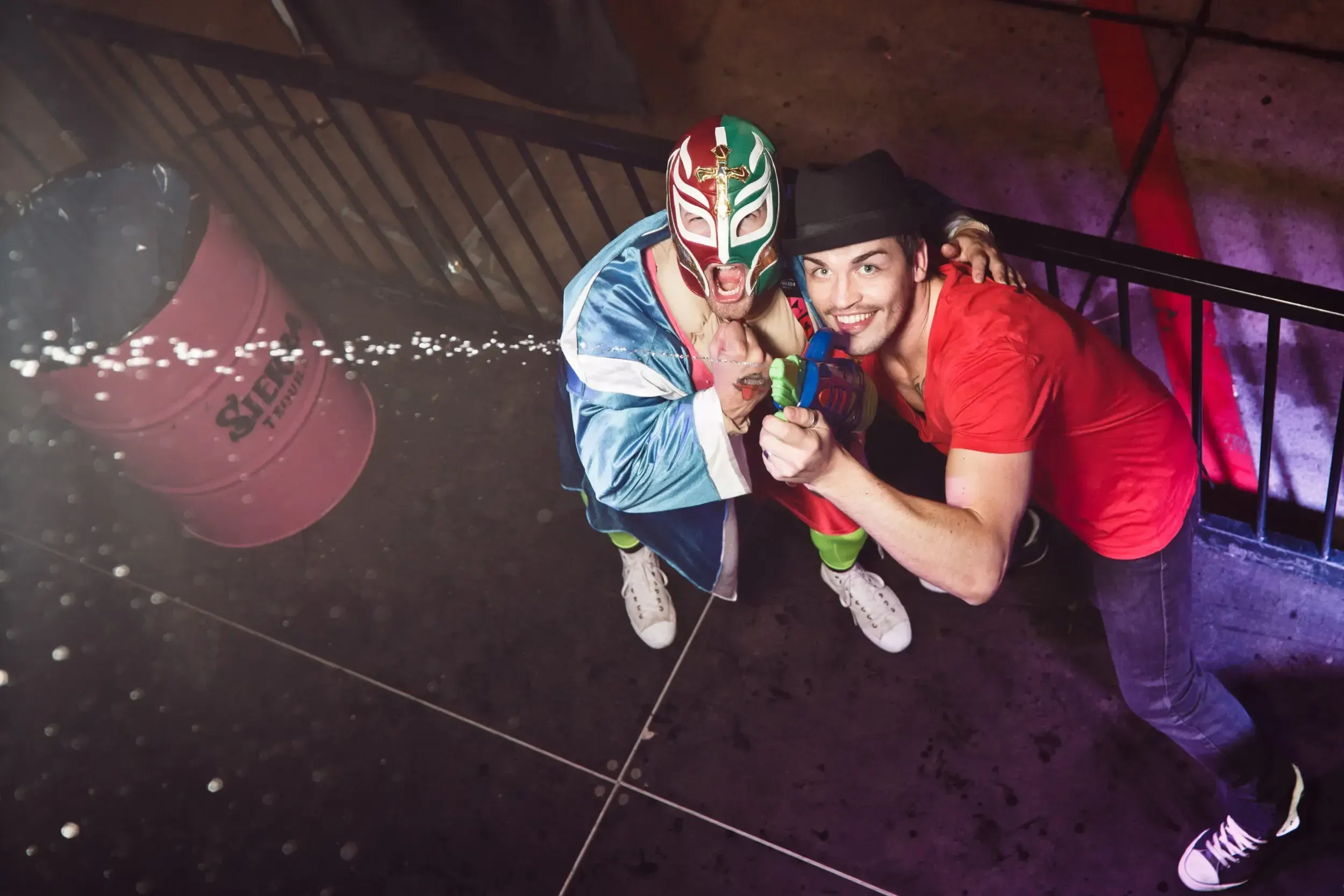Two friends posing on an outdoor staircase at night, one in a lucha libre mask spraying a water gun.