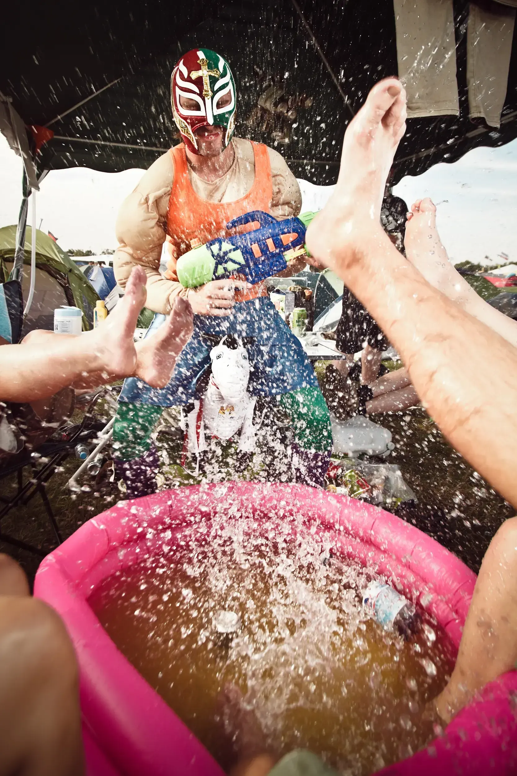 Man in a lucha libre wrestling mask spraying a water gun over a pink paddling pool at a festival campsite.