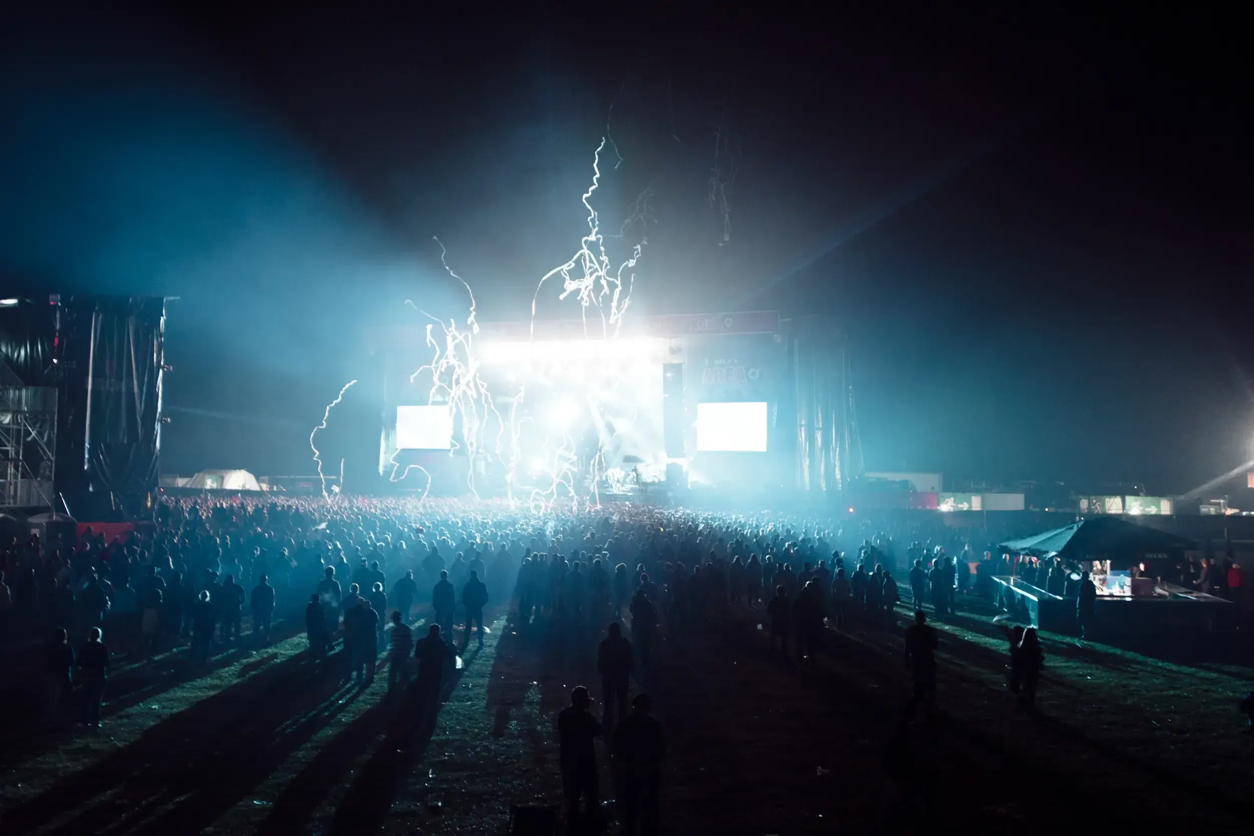 Crowd at a festival at night, backlit by bright stage lights with confetti streamers shooting into the air above the stage.
