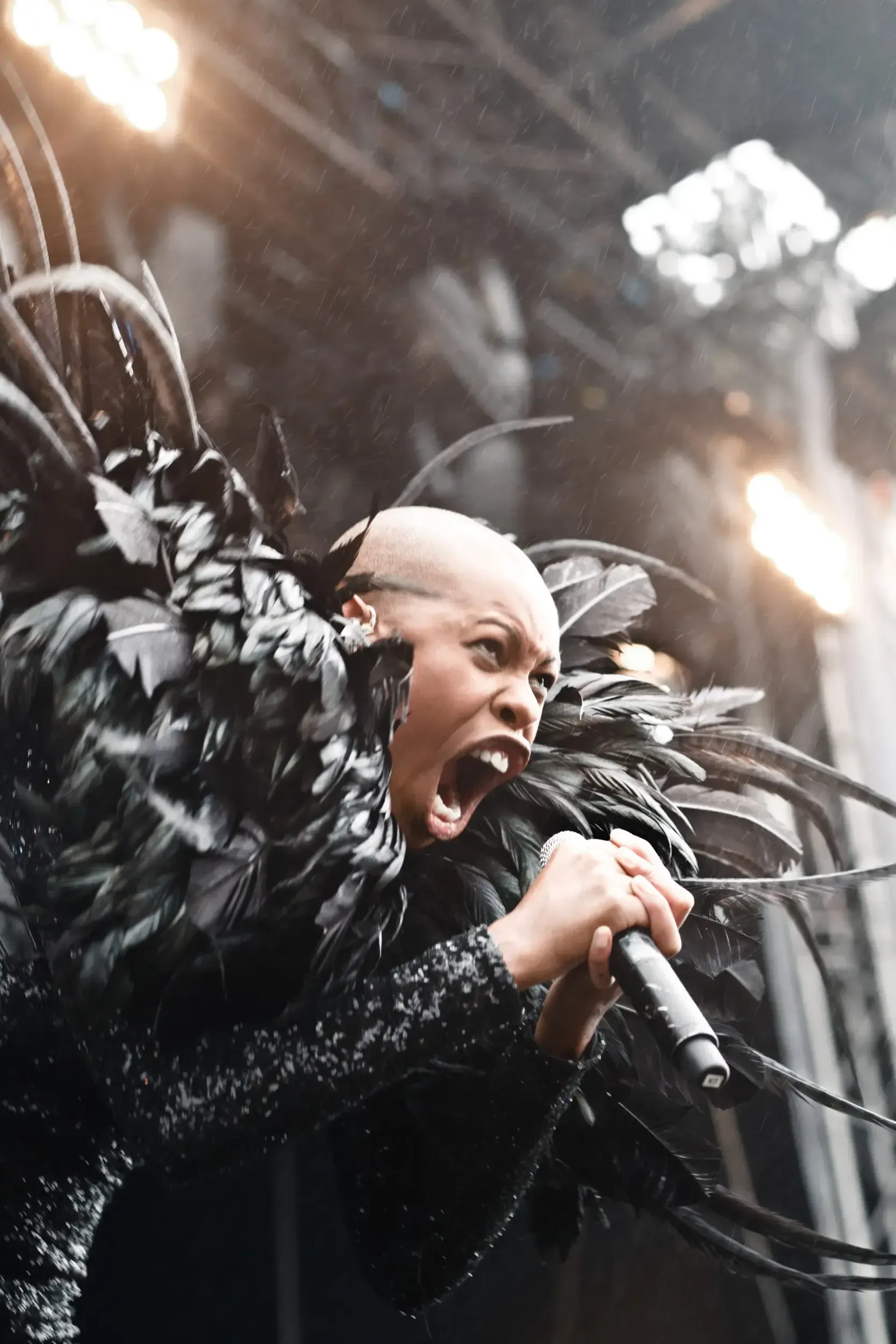 Rock singer with a shaved head and dramatic black feathered costume screaming into a microphone on a rain-soaked stage.