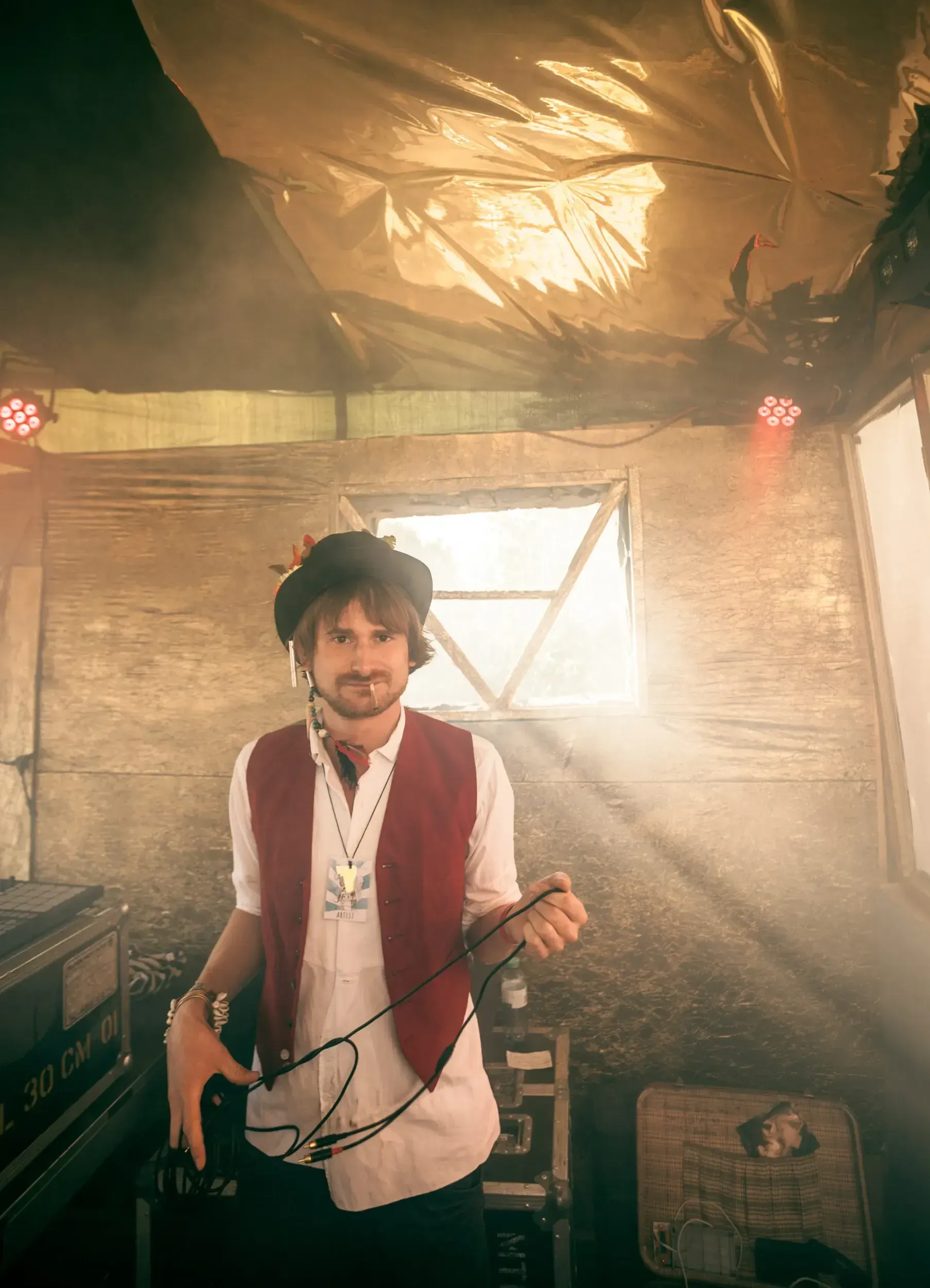 Man in a top hat and red waistcoat standing in a sunlit backstage tent holding cables.