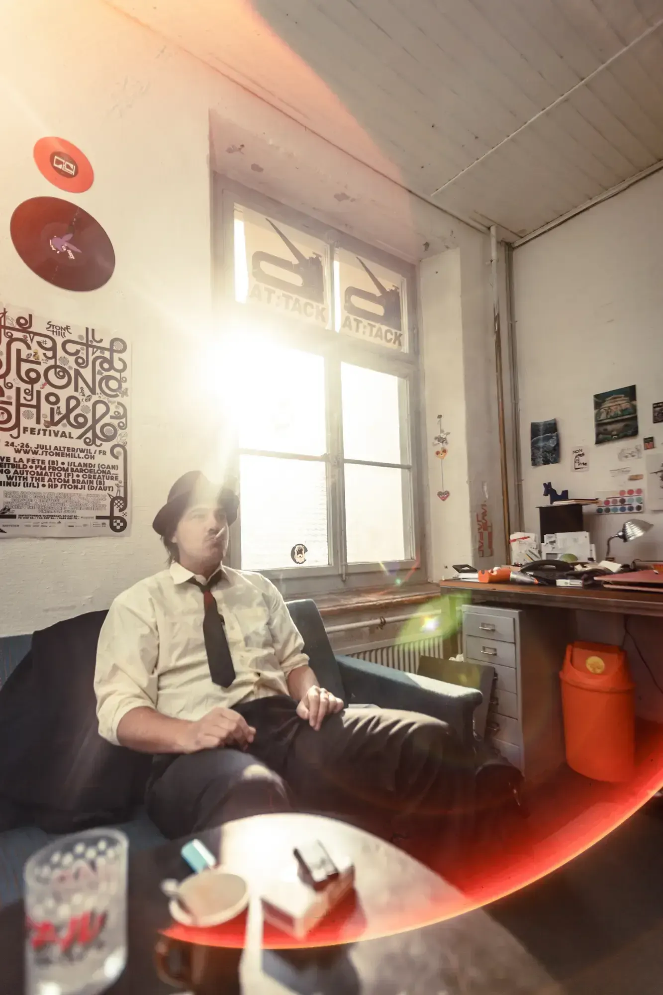Man in a shirt and tie sitting in an armchair in a sunlit studio decorated with band posters.