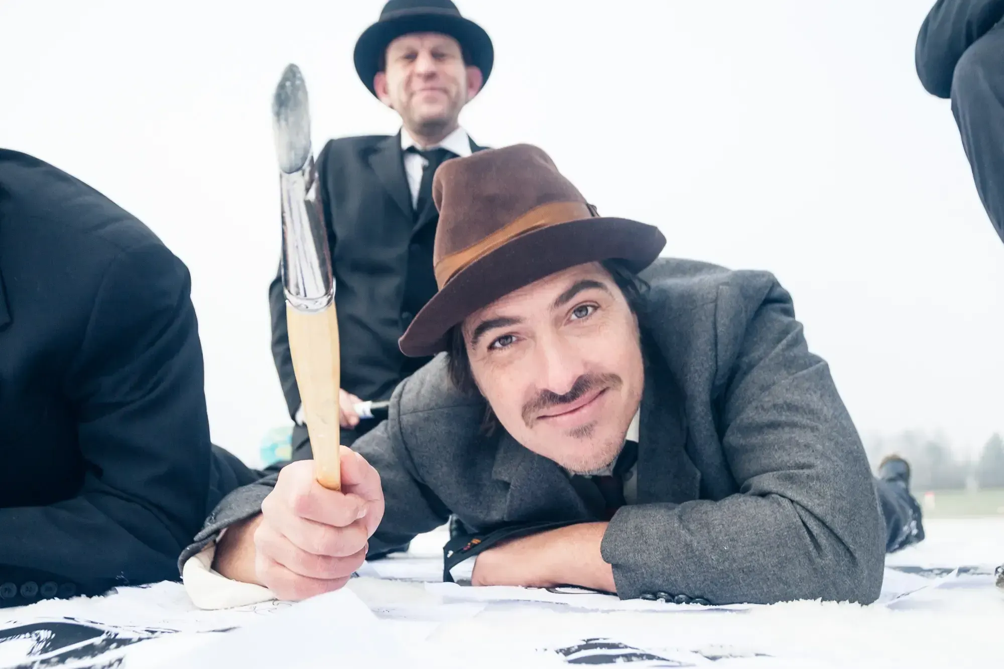 Close-up of a Büro Destruct designer in a fedora holding up a large paintbrush while lying over sketch sheets.