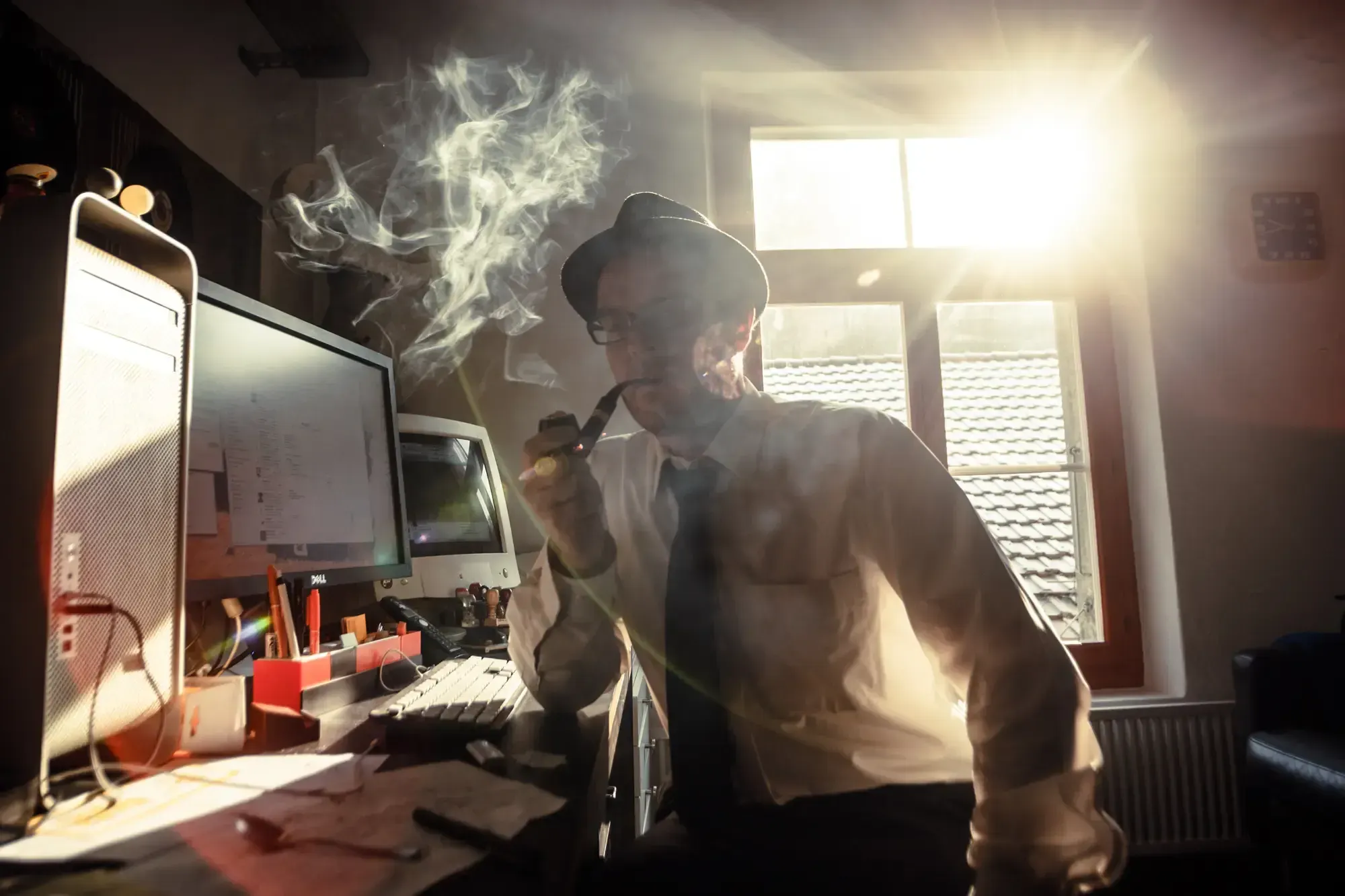 Man in a hat and glasses smoking a pipe at a desk with computer monitors, backlit by a window.