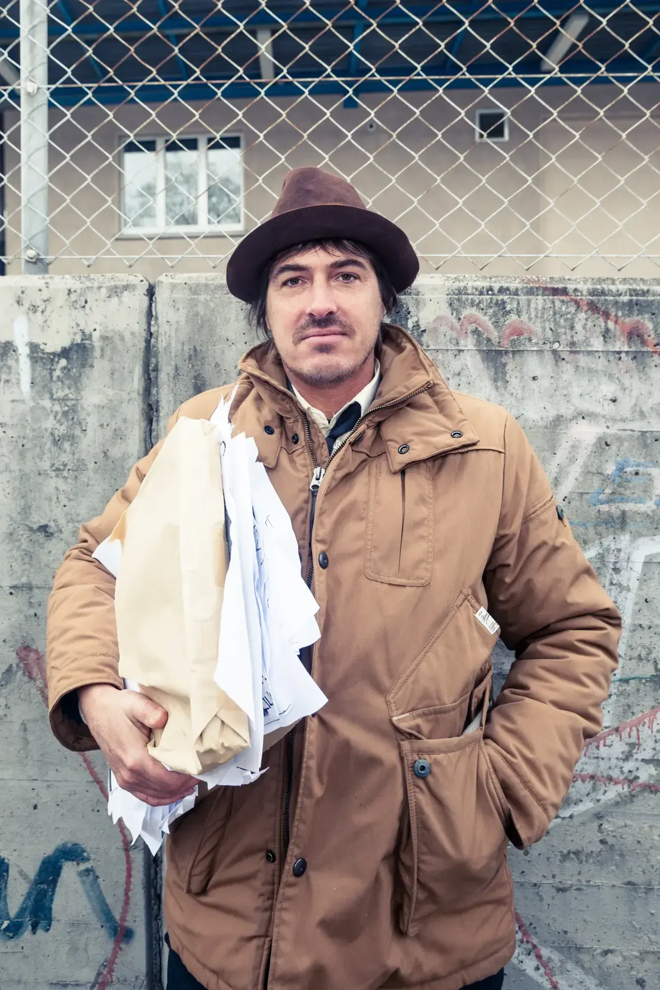 Portrait of a Büro Destruct designer in a hat and jacket holding a stack of papers in front of a graffitied concrete wall and chain-link fence.