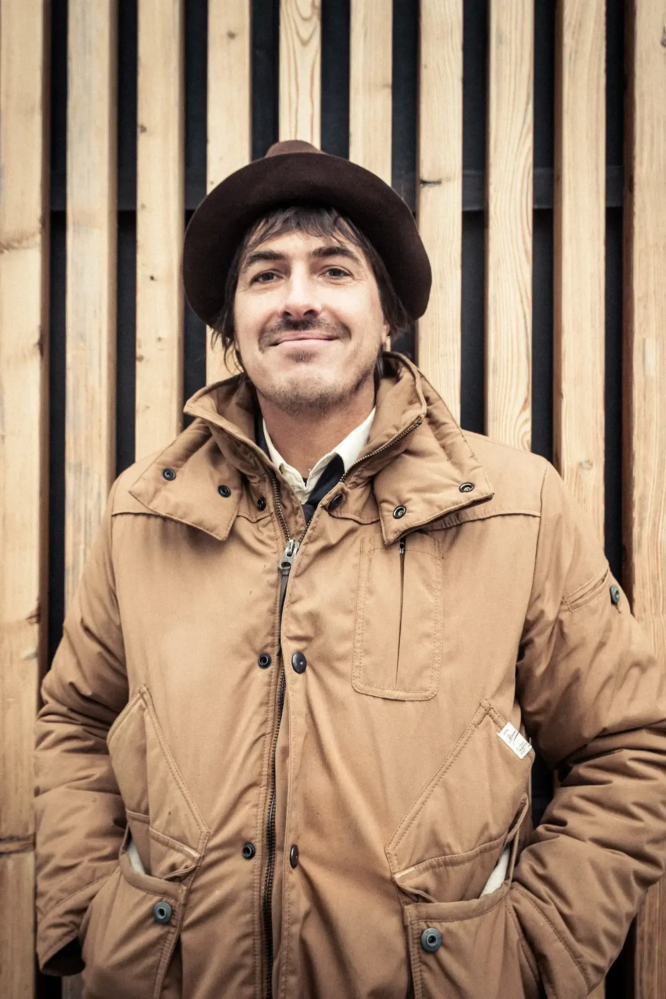 Smiling portrait of a Büro Destruct designer in a hat and jacket against a wooden slat wall.