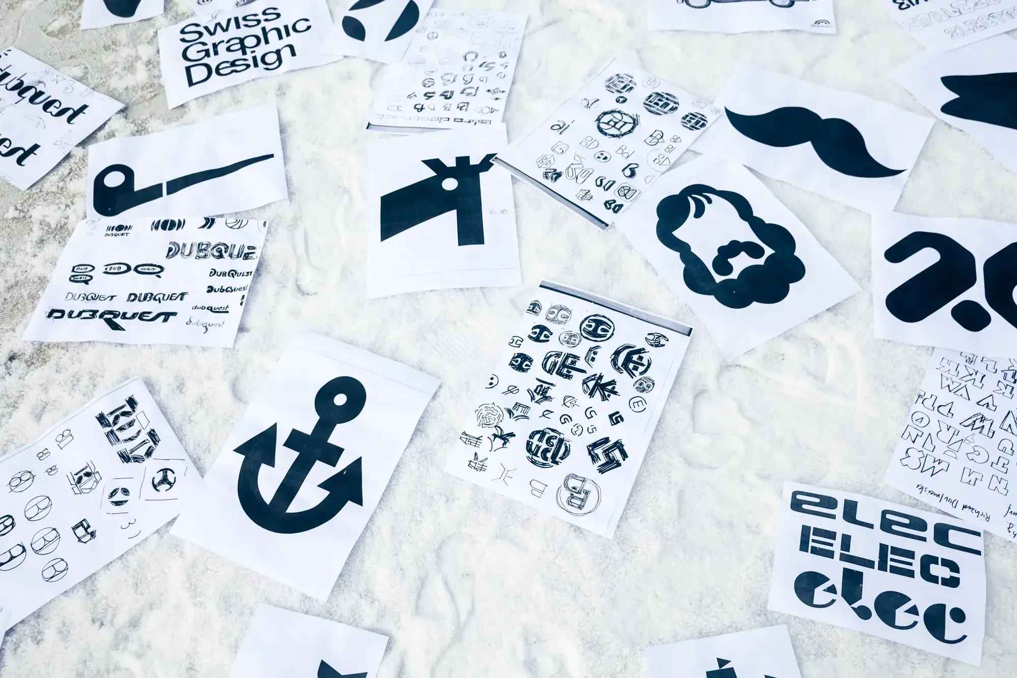 Sheets of hand-drawn logo sketches and lettering studies laid out on white sand, including a 'Swiss Graphic Design' sign.
