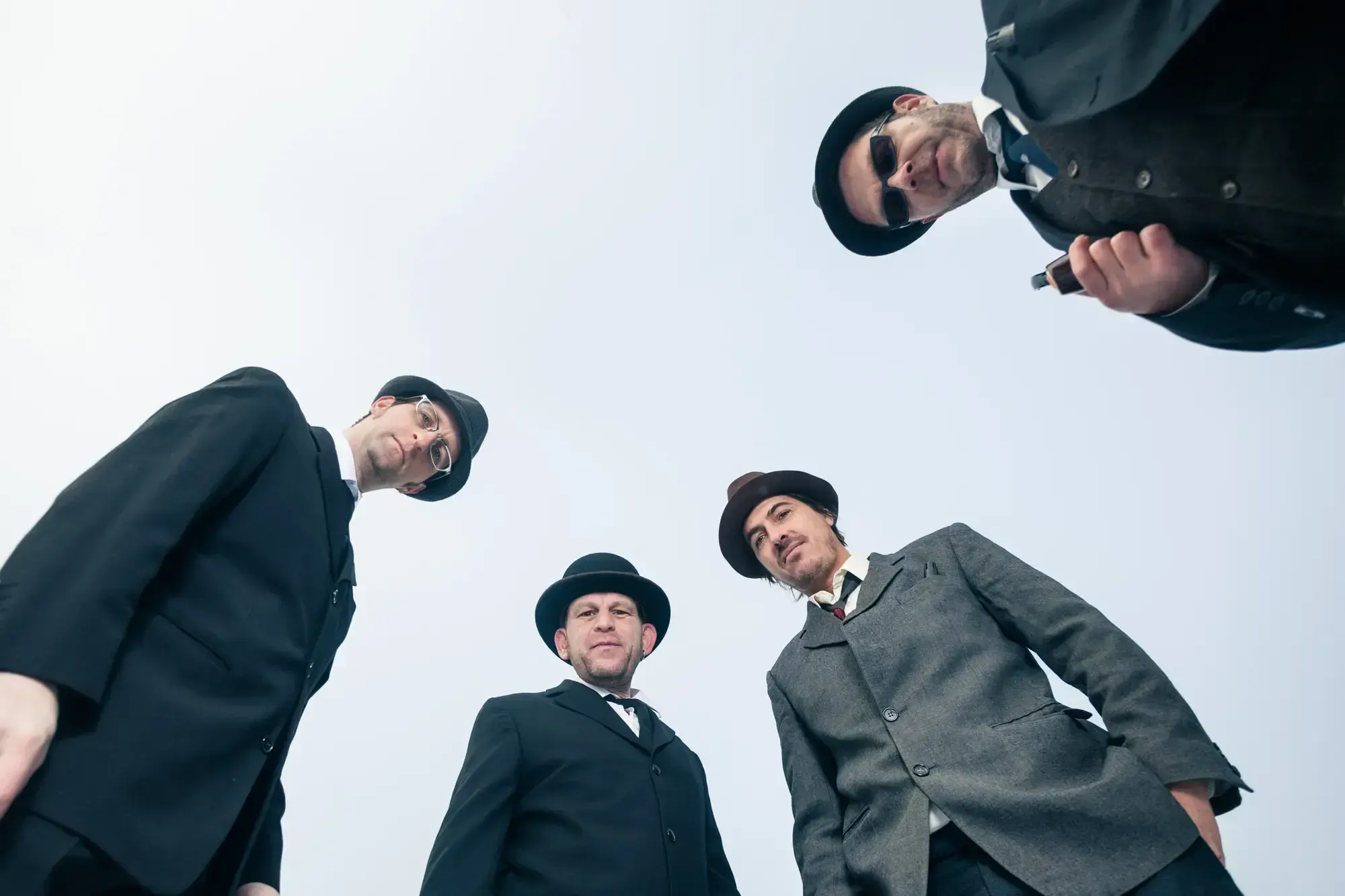 Low-angle portrait of the Büro Destruct team in hats and suits looking down at the camera against a bright sky.
