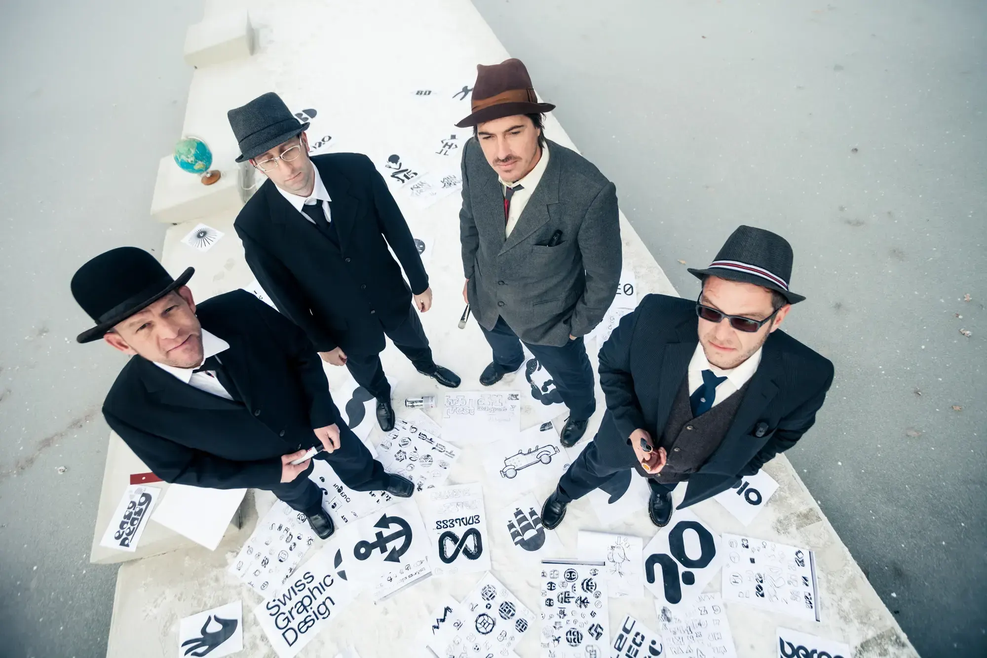 Overhead view of the Büro Destruct team in hats standing among sheets of hand-drawn logo sketches, including a 'Swiss Graphic Design' sign.