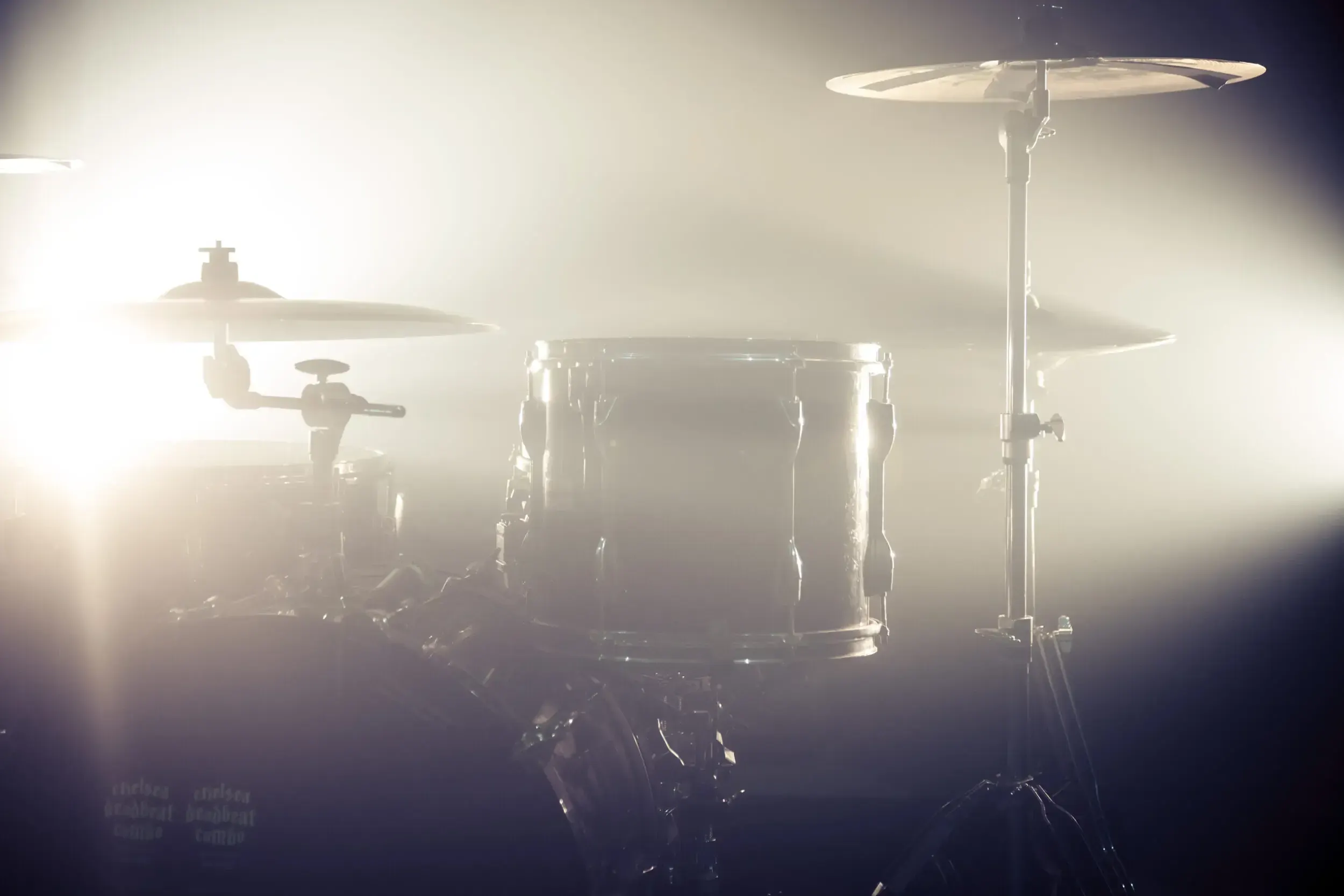Drum kit silhouetted against bright stage lights and haze.