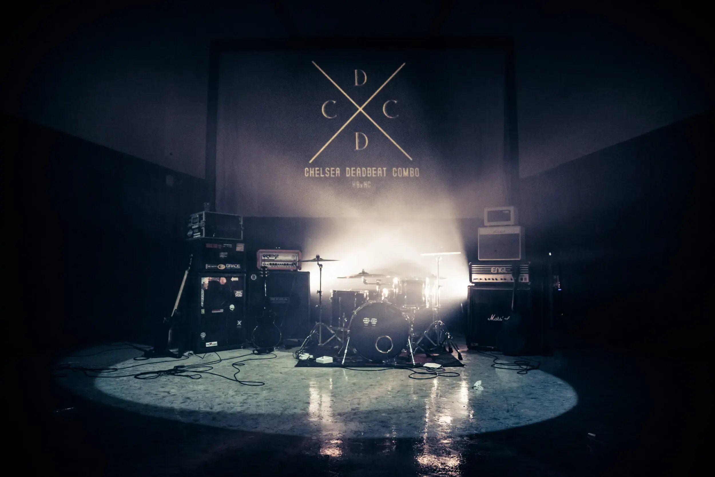 Empty stage set up with a drum kit and amplifiers beneath a projected Chelsea Deadbeat Combo logo.