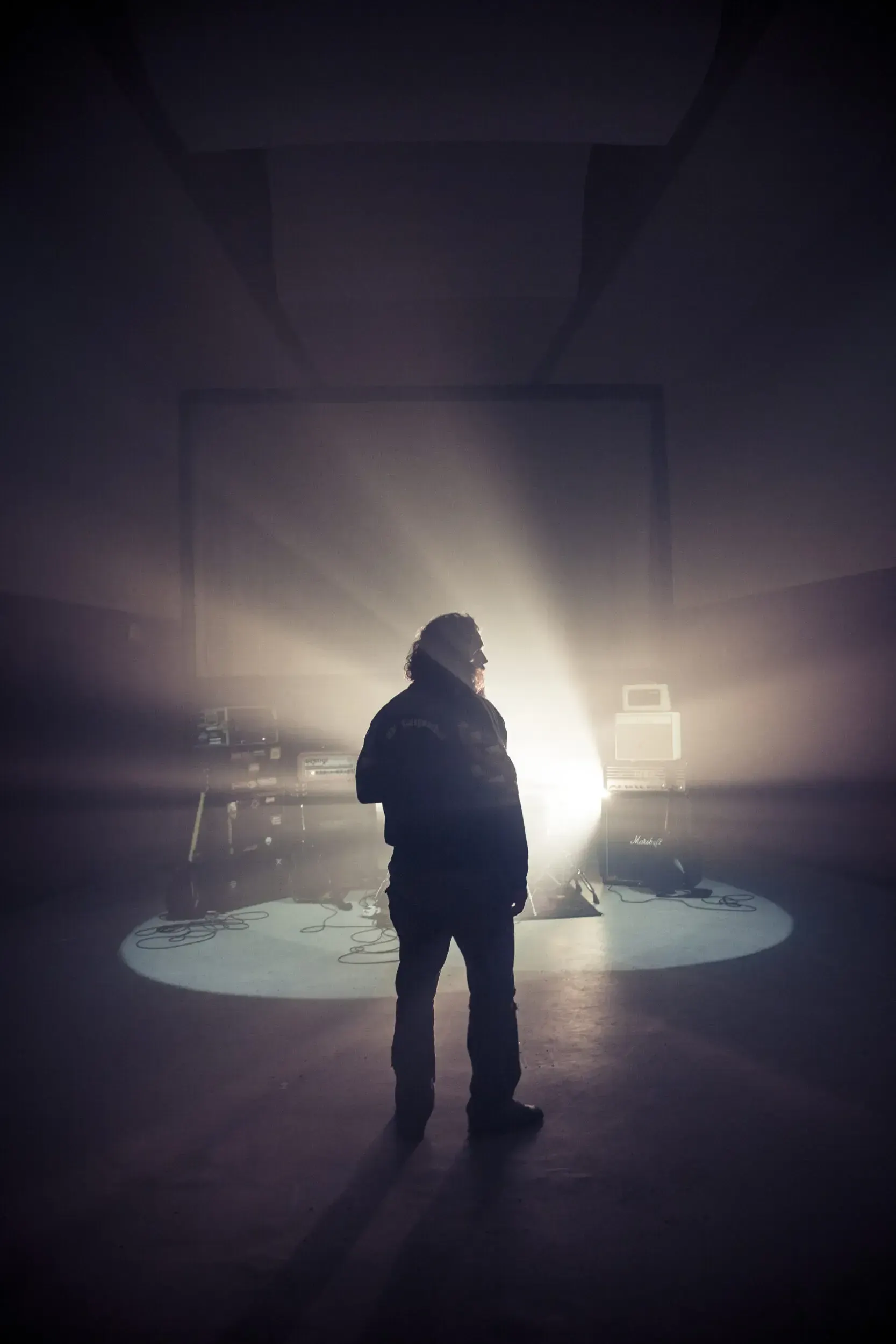 Silhouette of a musician standing in a spotlight on an empty stage surrounded by amplifiers.