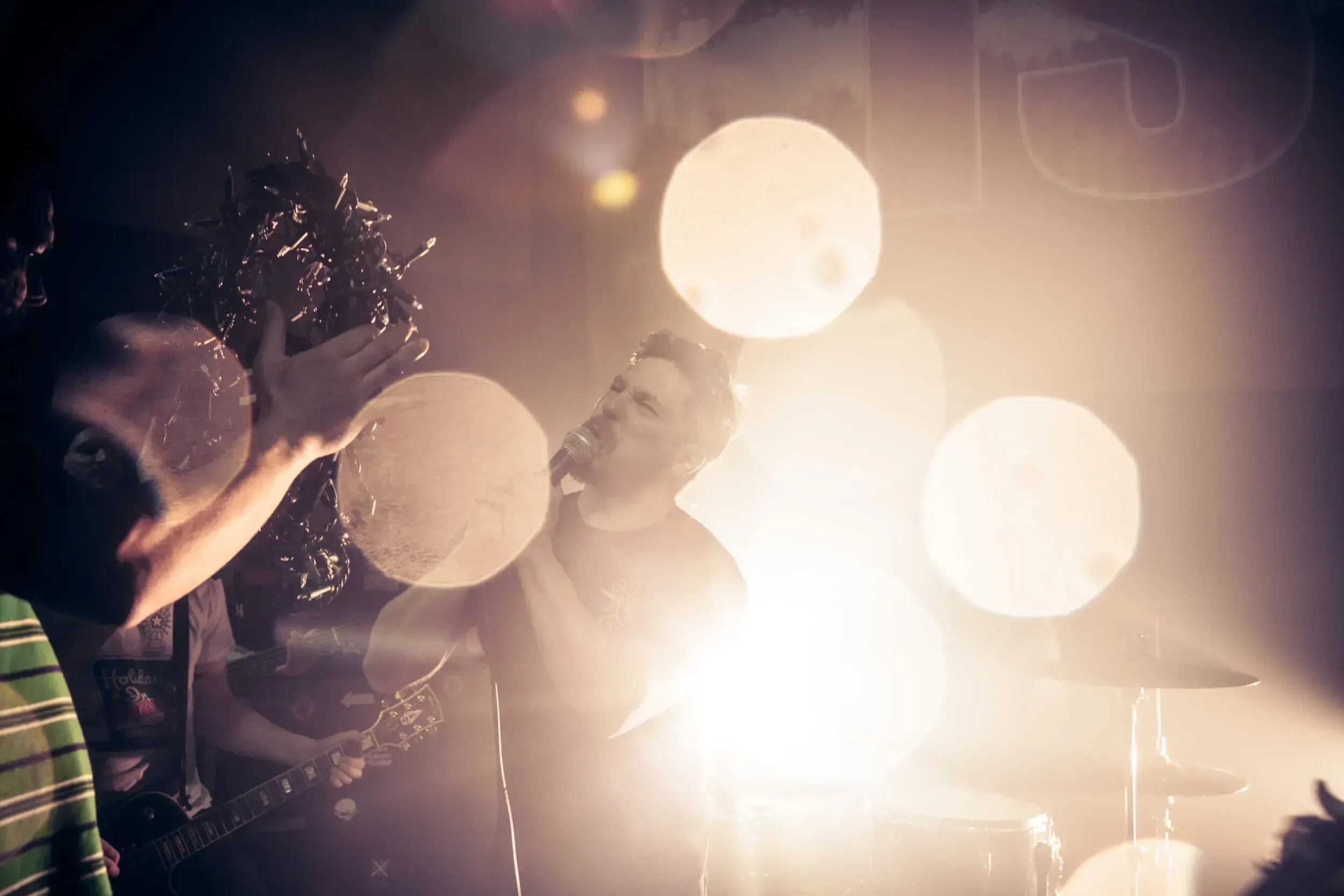 Singer performing into a microphone, backlit by bright stage lights with the crowd's hand visible in the foreground.