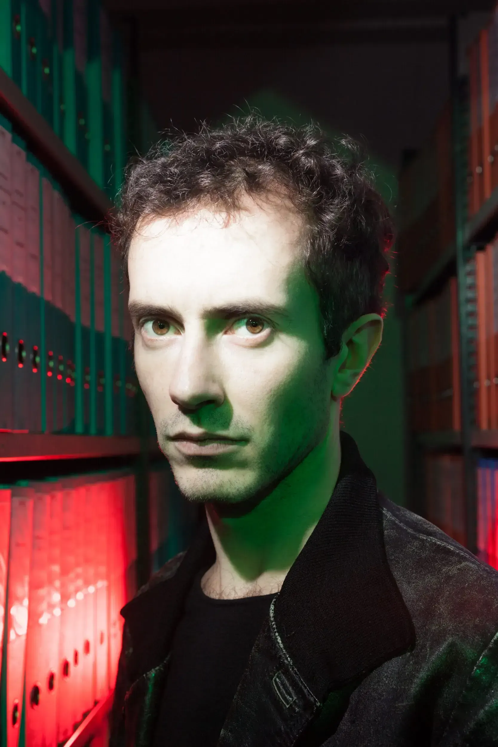 Portrait of Claudius Körber lit in red and green standing among file-lined archive shelves.