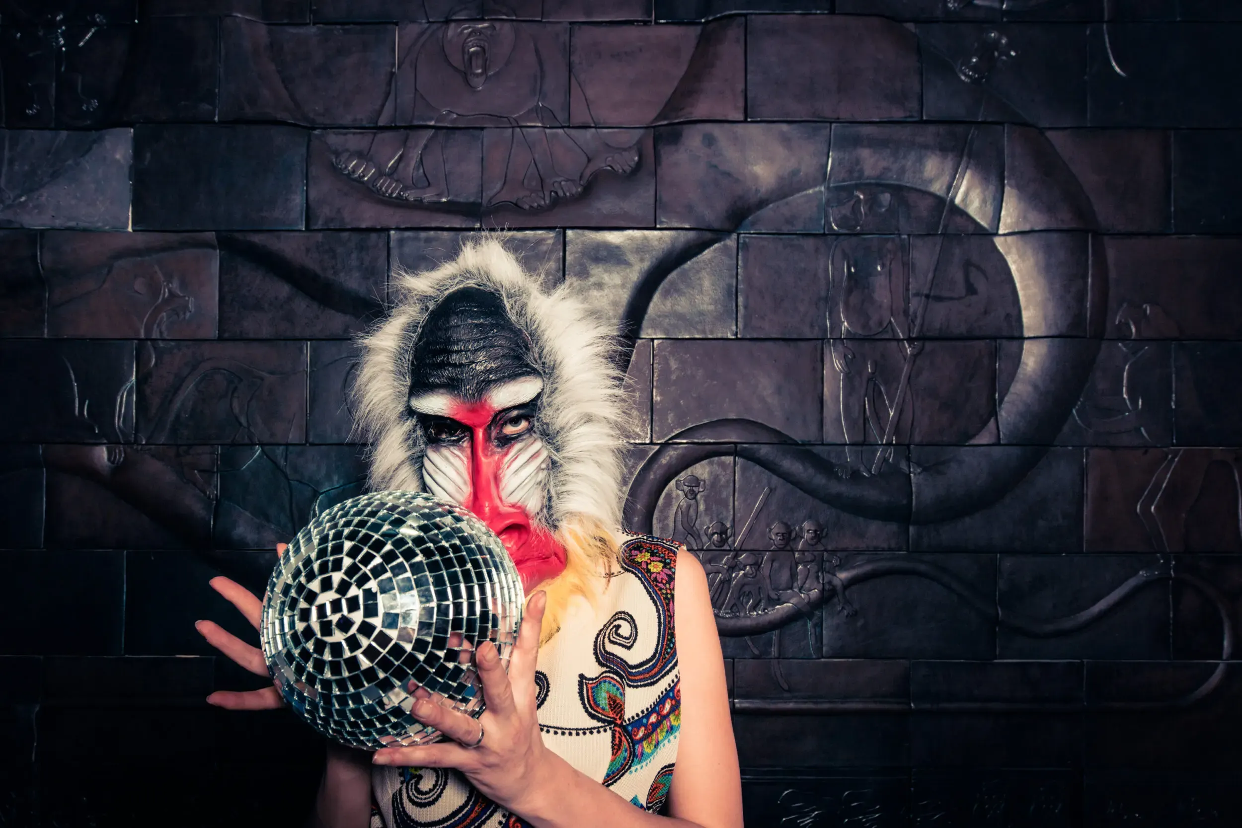 Guest in a baboon mask holding up a disco ball against a graffiti-covered wall.