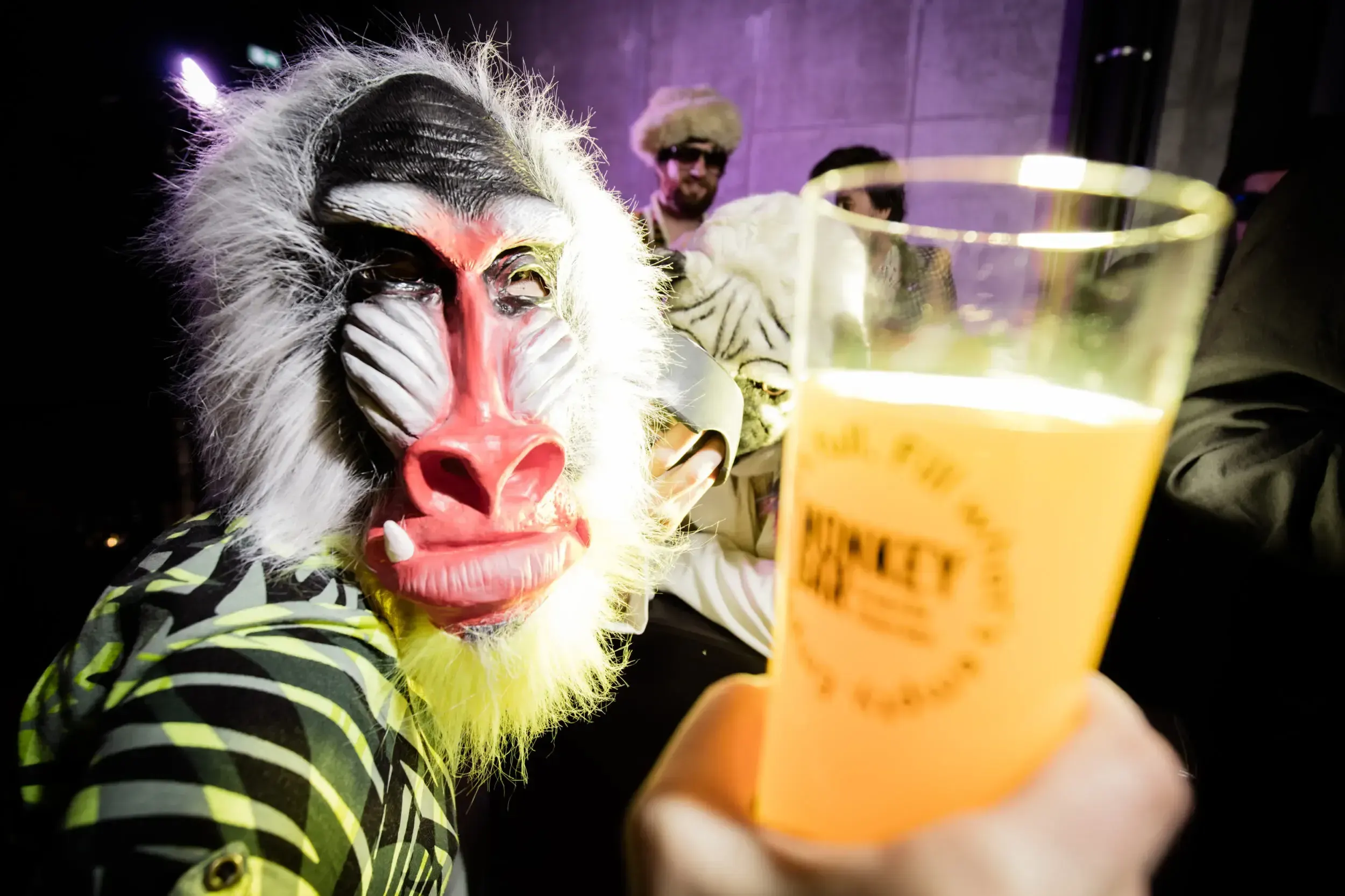 Close-up of a guest in a baboon mask holding up a drink to the camera.