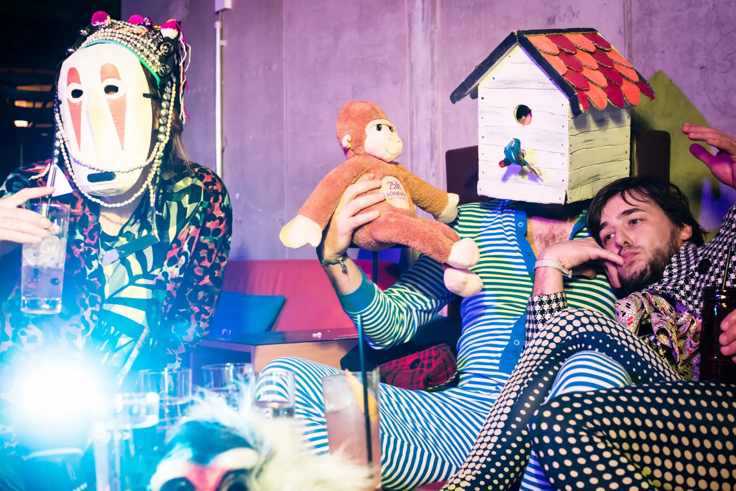 Guests in a birdhouse mask and a painted face mask relaxing together with a toy monkey.