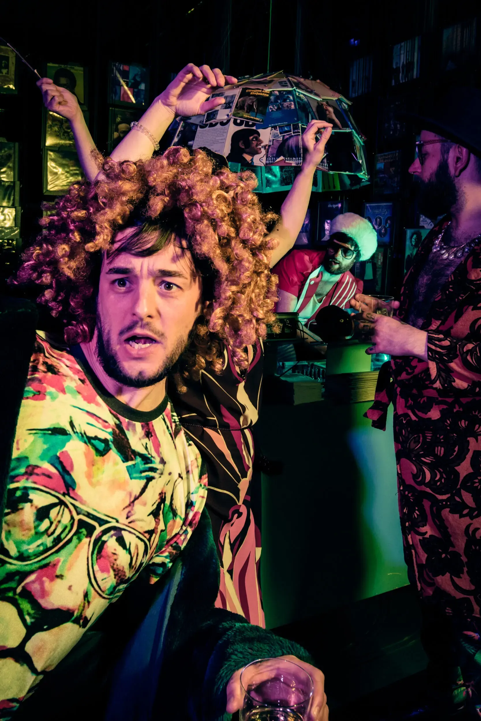 Man in a curly wig and animal-print jacket raising his arms among other costumed guests.