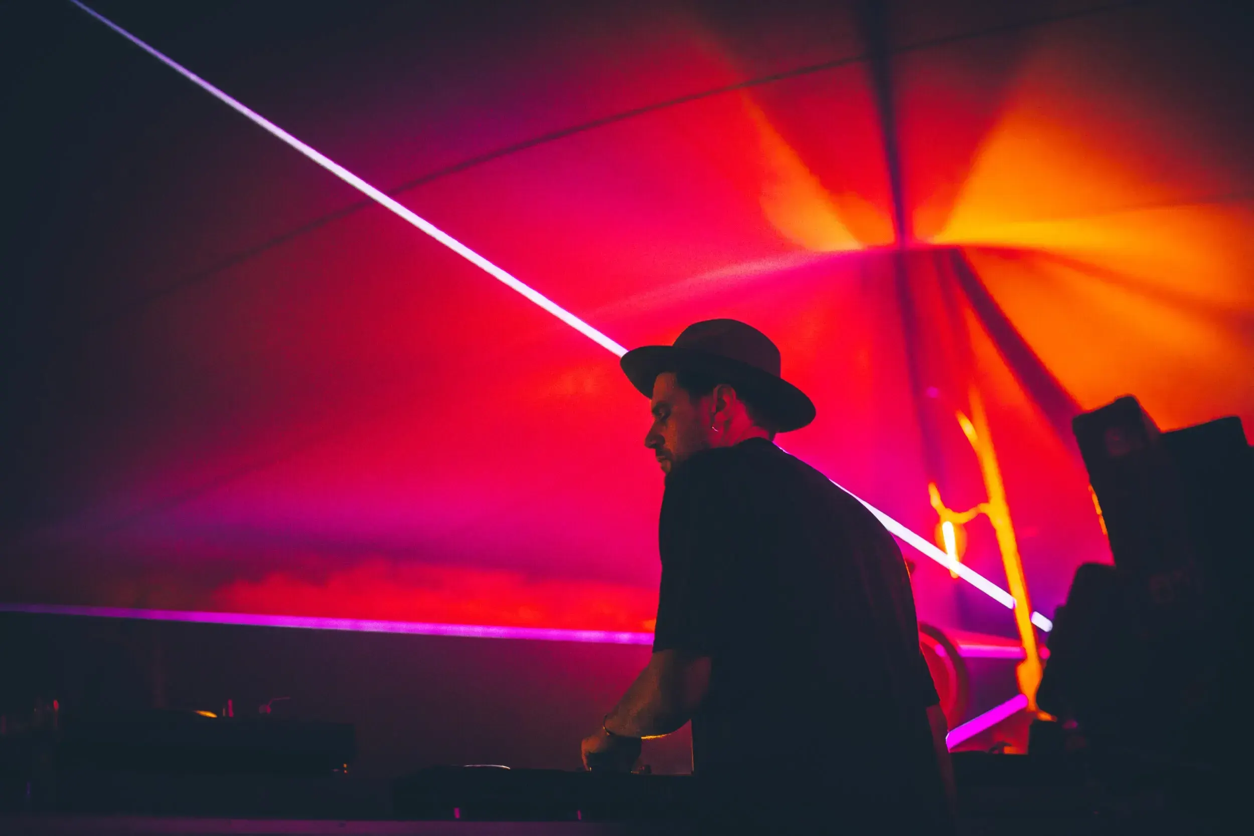 Silhouette of a DJ in a hat under red and orange laser lights at a costume party.