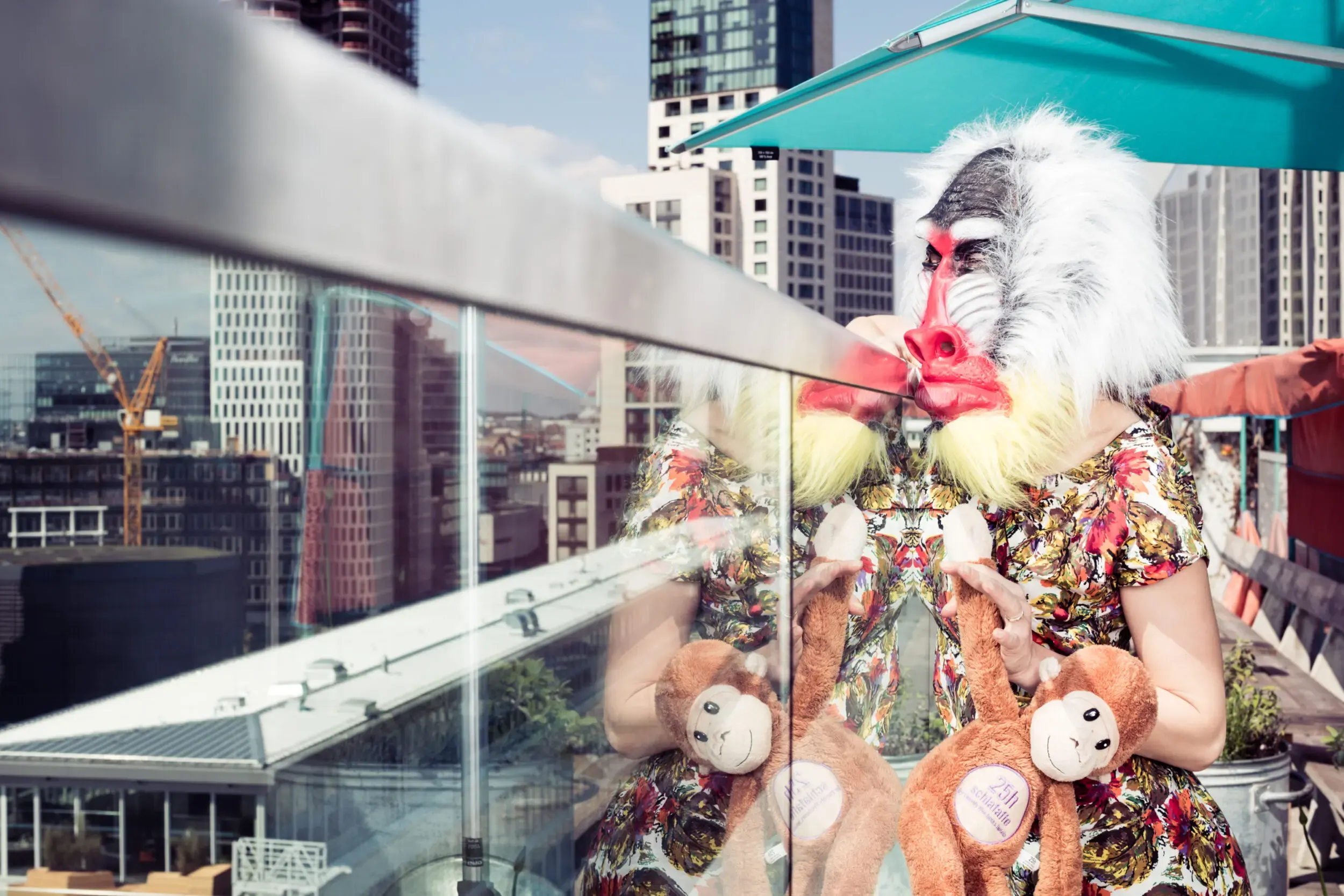 Guest in a feathered mask on a rooftop terrace beneath an umbrella, skyline behind.