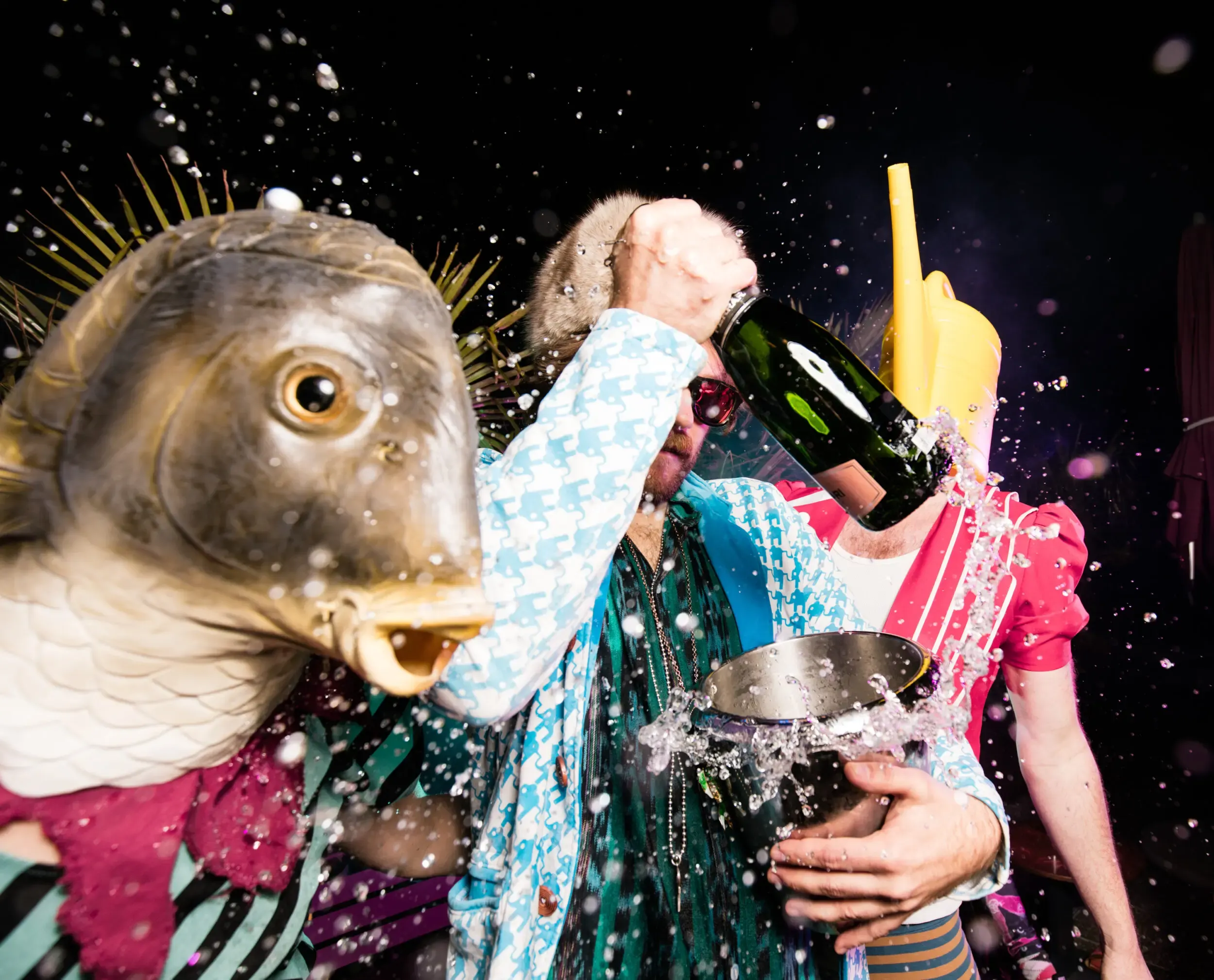 Guest in a fish mask amid others pouring champagne into a metal bowl, water splashing everywhere.